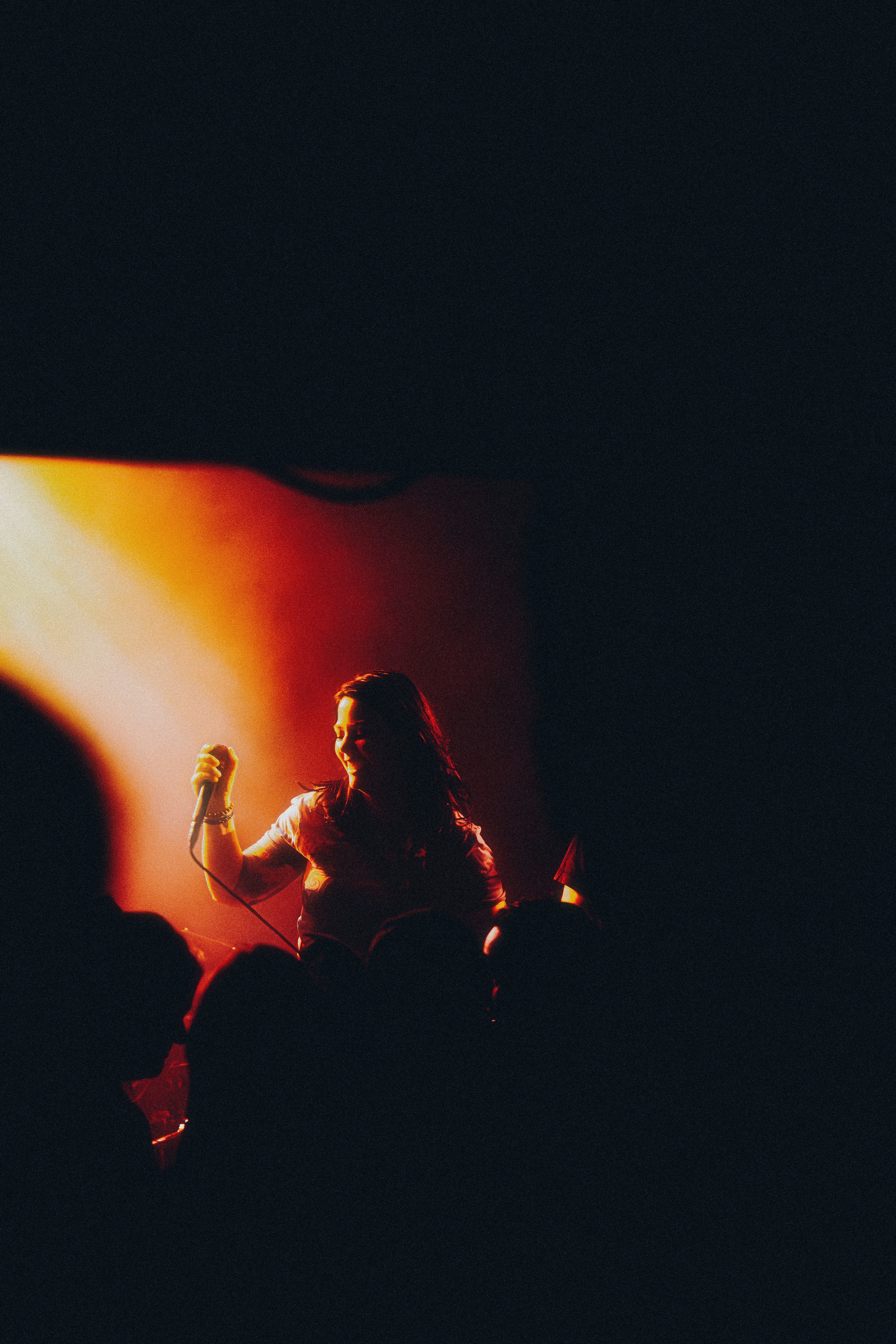 A woman holding a microphone on stage, silhouetted against warm orange and red lighting, with audience members in the foreground.