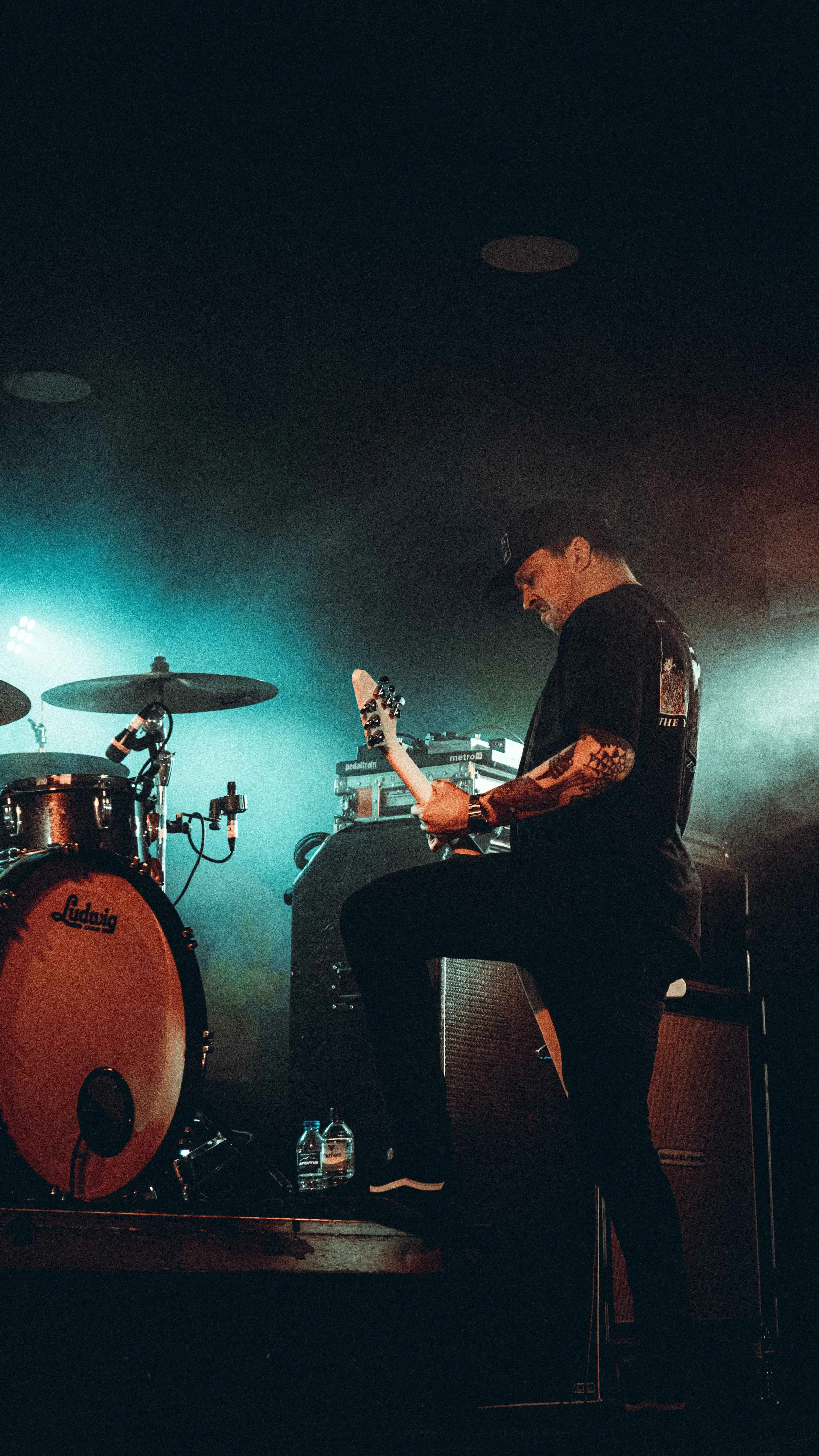 A musician playing an electric guitar on stage with stage lights and sound equipment in the background.