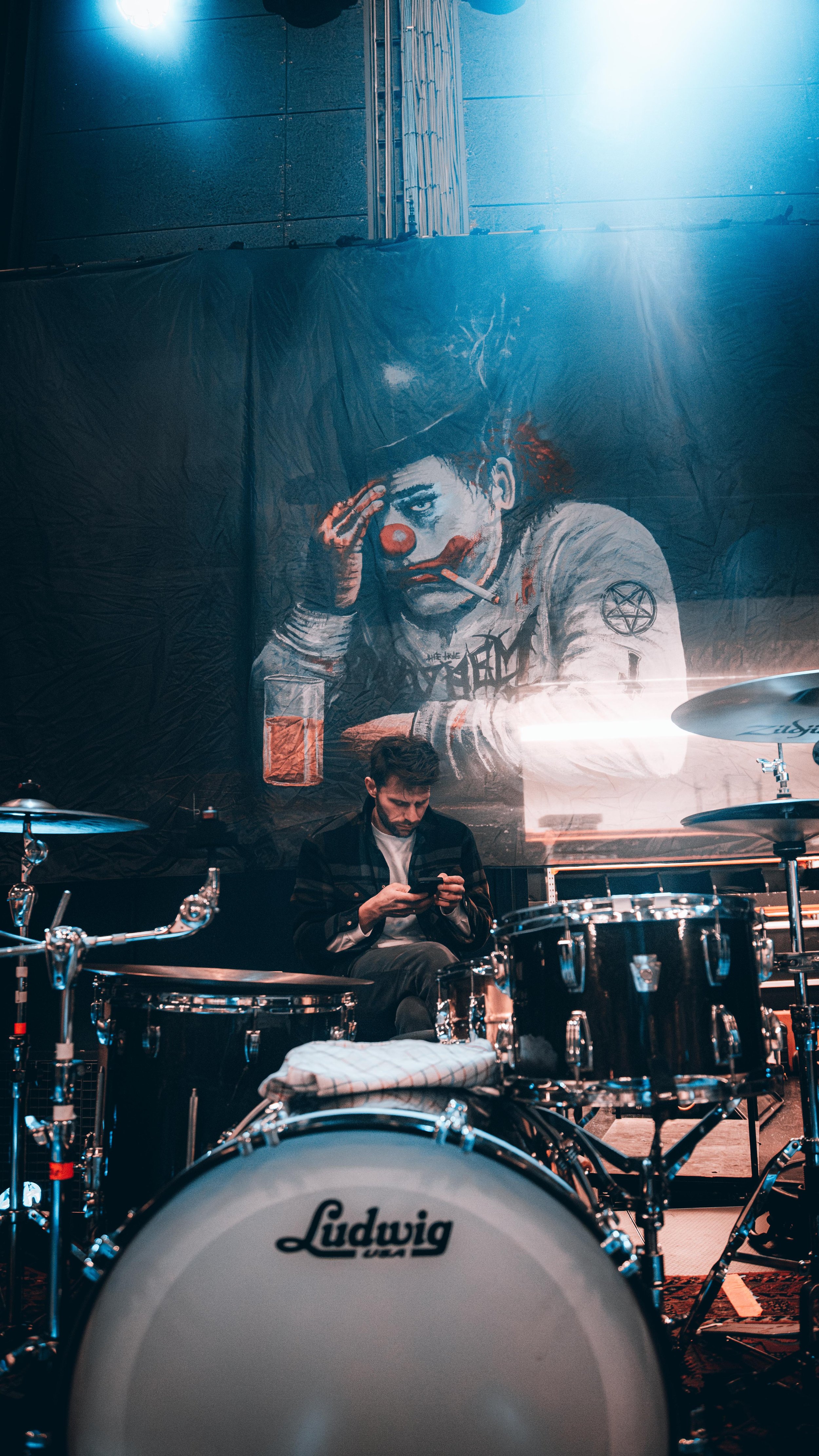 A man sitting behind a Ludwig drum set in a room with a large graffiti mural of a clown with a cigarette, a glass of orange juice, and tattoos on his arm in the background.