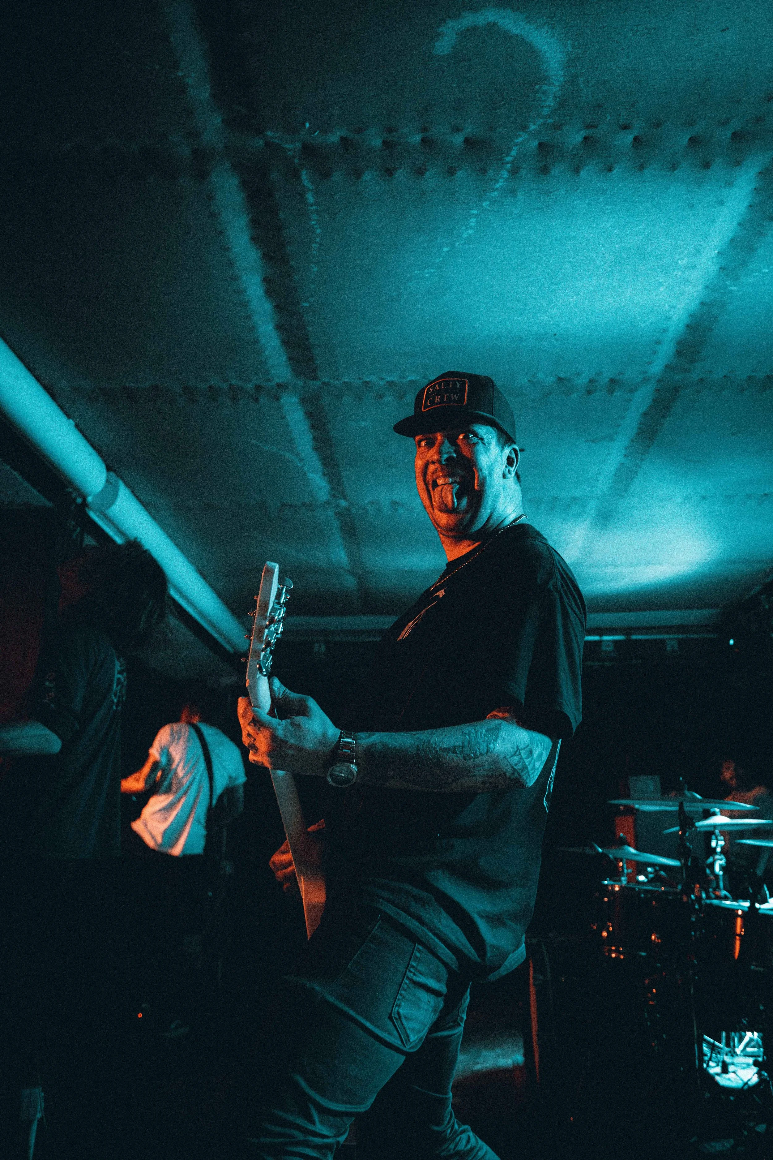 A man in a black cap and T-shirt making a silly face while playing an electric guitar during a live music performance at a nightclub or concert venue. The background includes other musicians and blue stage lighting.