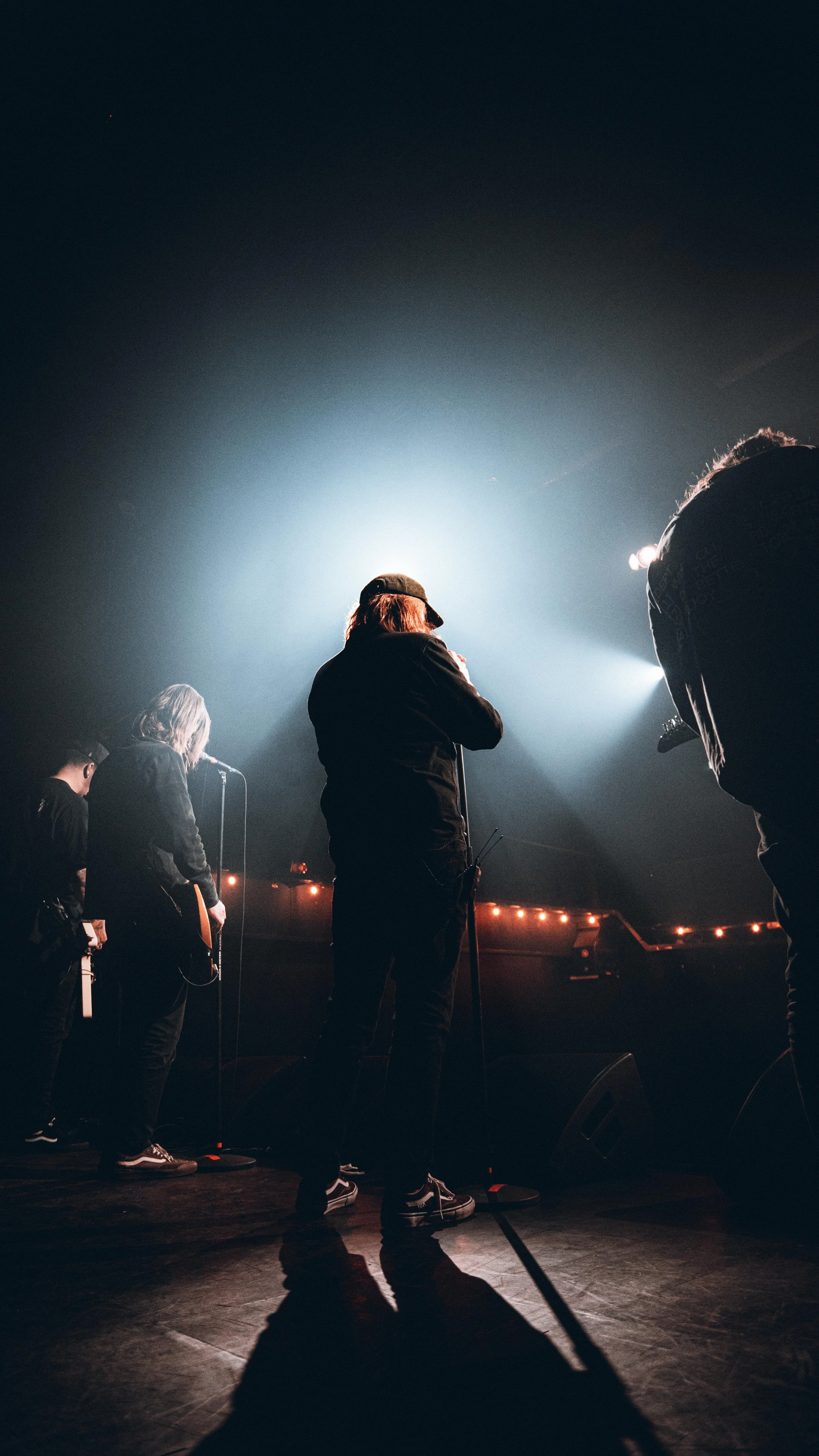 Musicians performing on stage with backlighting, featuring a person in the center with a microphone and others playing instruments.