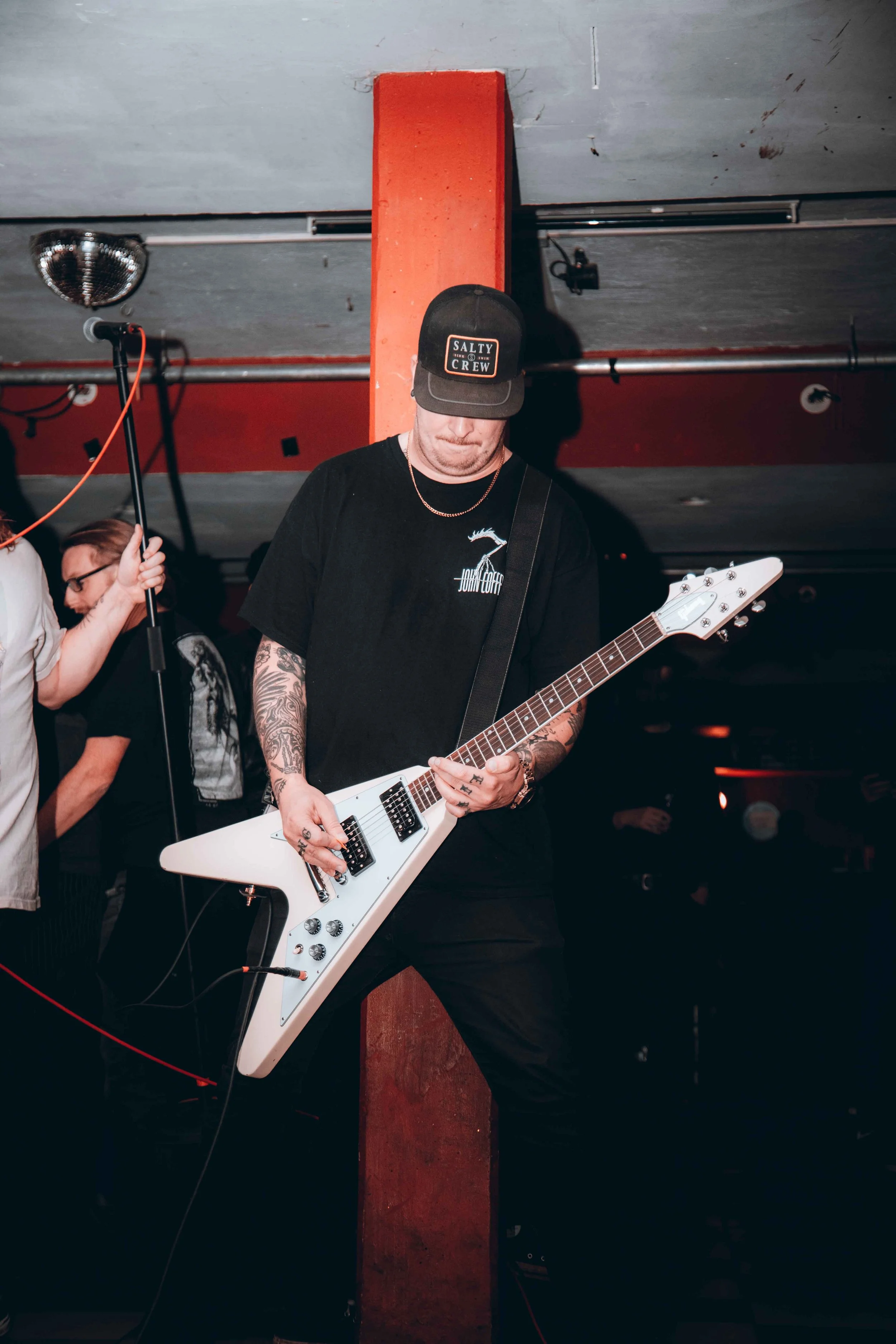 A musician playing a white electric guitar in a dimly lit bar or club, wearing a black cap and T-shirt, with tattoos on his arms.