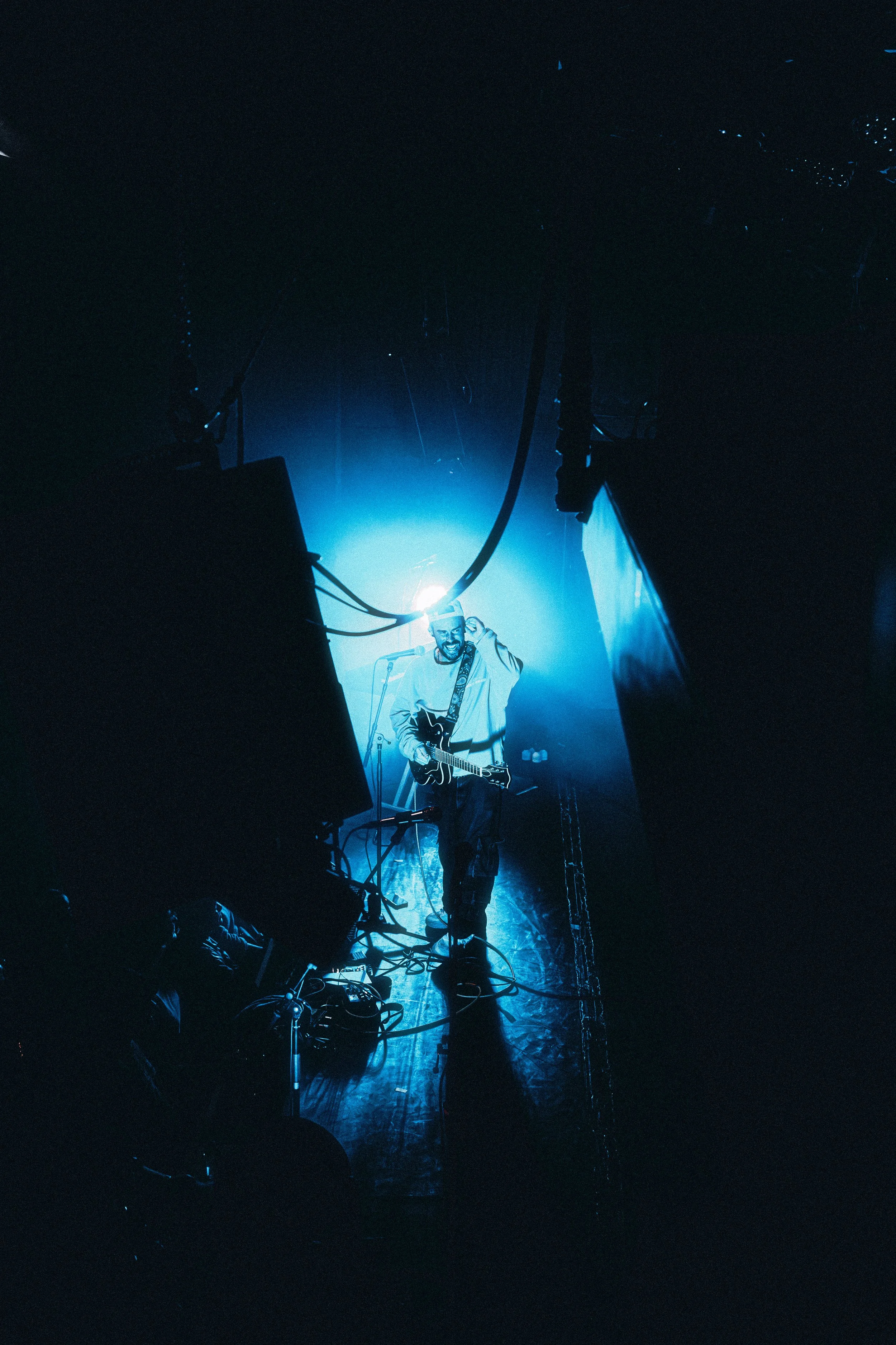A musician performing on stage with a guitar, illuminated by blue stage lighting, surrounded by equipment and cables, viewed from a low angle.