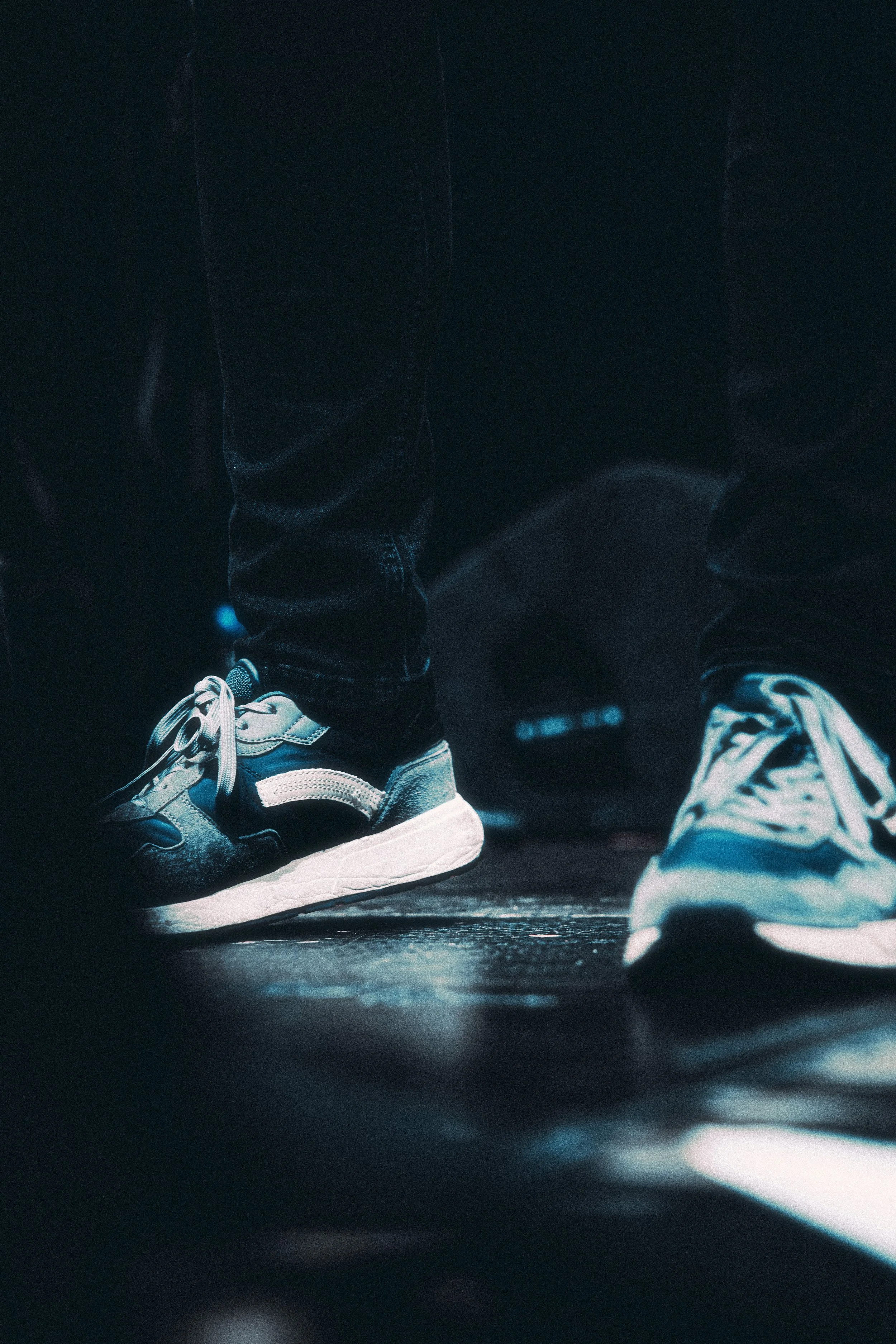 Close-up of a person's feet wearing sneakers and black jeans, stepping on a dark floor with a highlighted reflection, in a dimly lit environment.