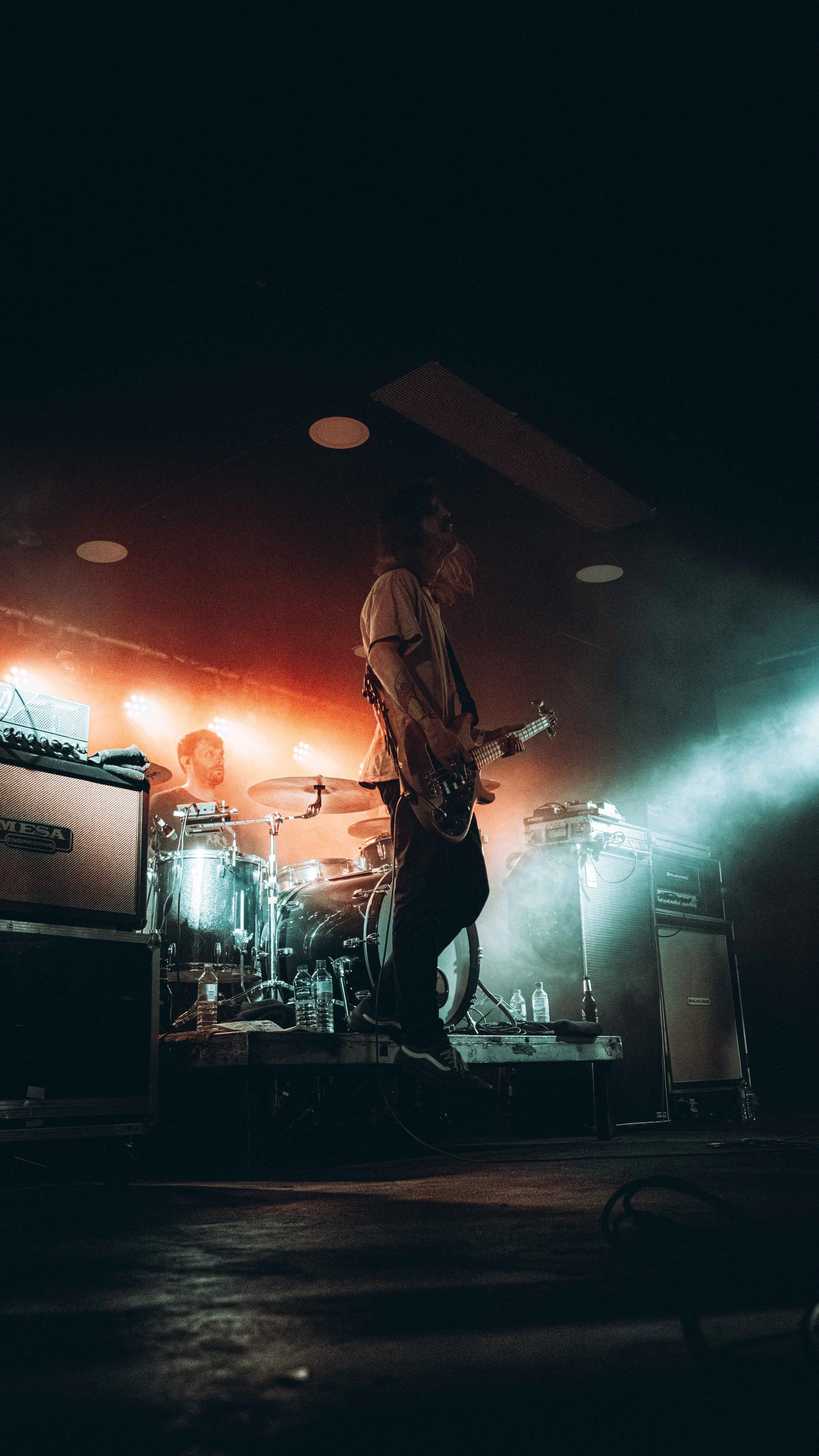 Musicians performing on stage, with a guitarist in the foreground and a drummer in the background, illuminated by stage lighting.