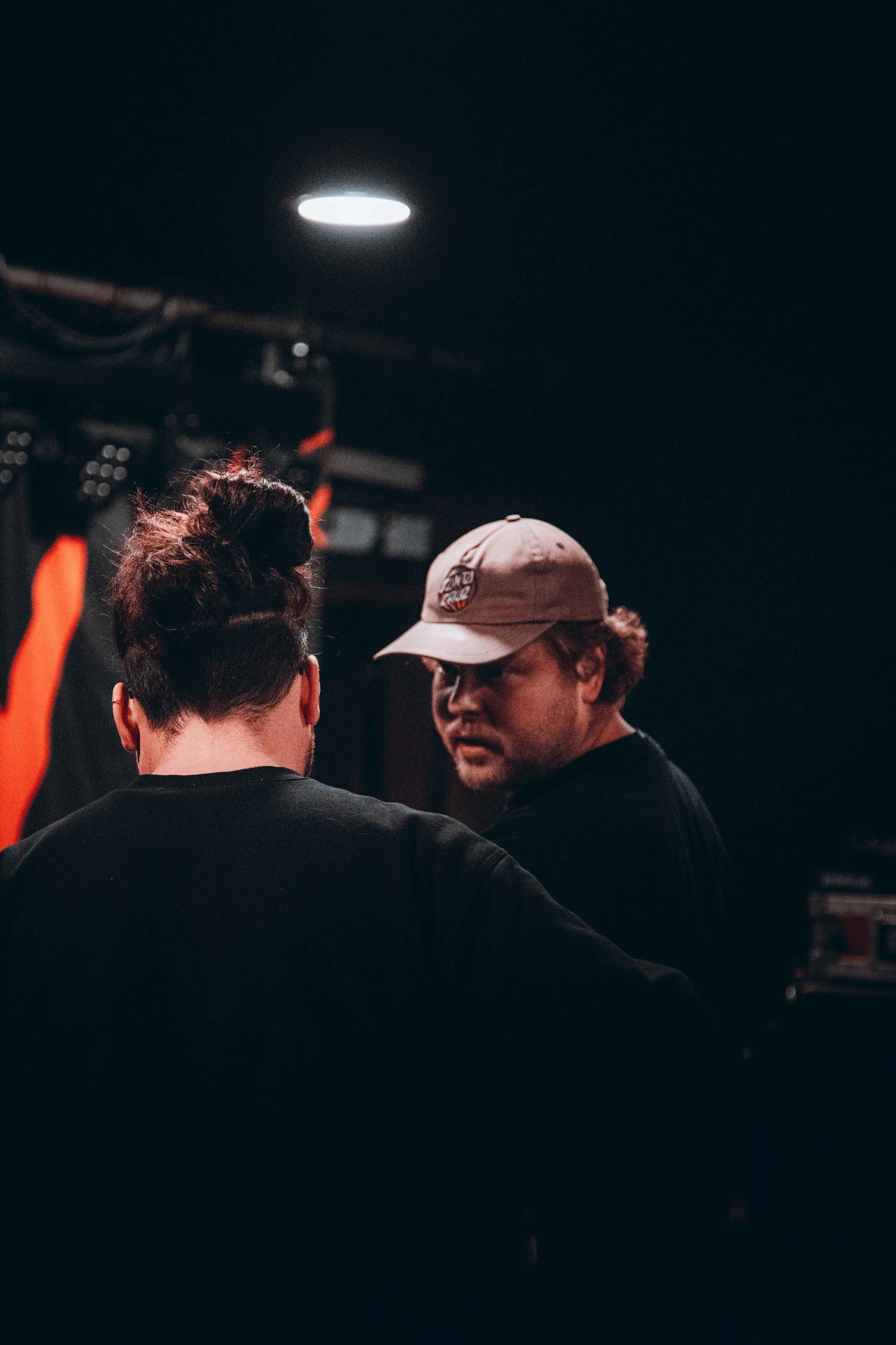 Two people in conversation in a dark room, one facing away with a man wearing a cap, and the other partially visible.