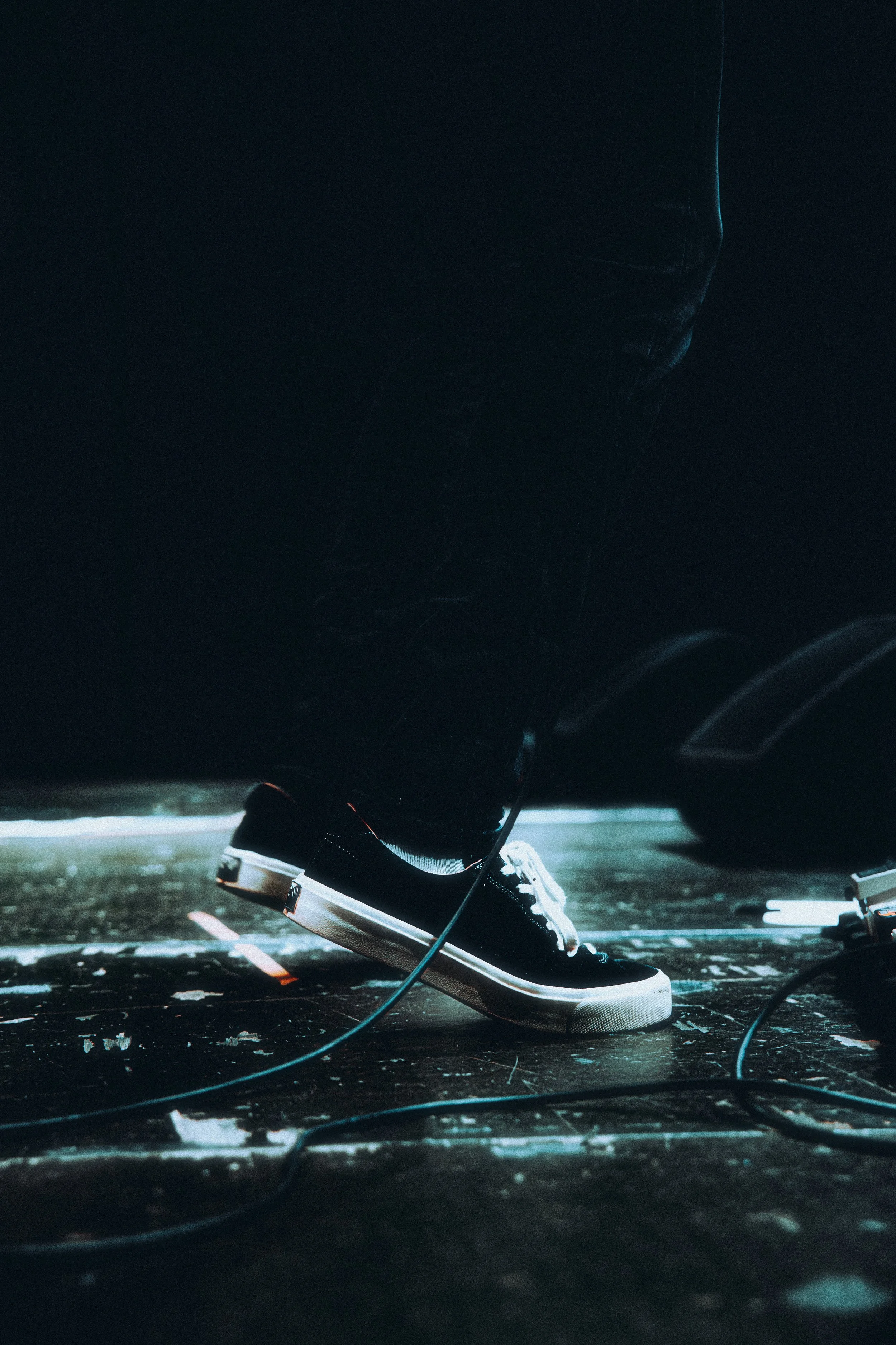 Close-up of a person wearing black sneakers with white laces standing on a dark stage floor, with wires scattered around.