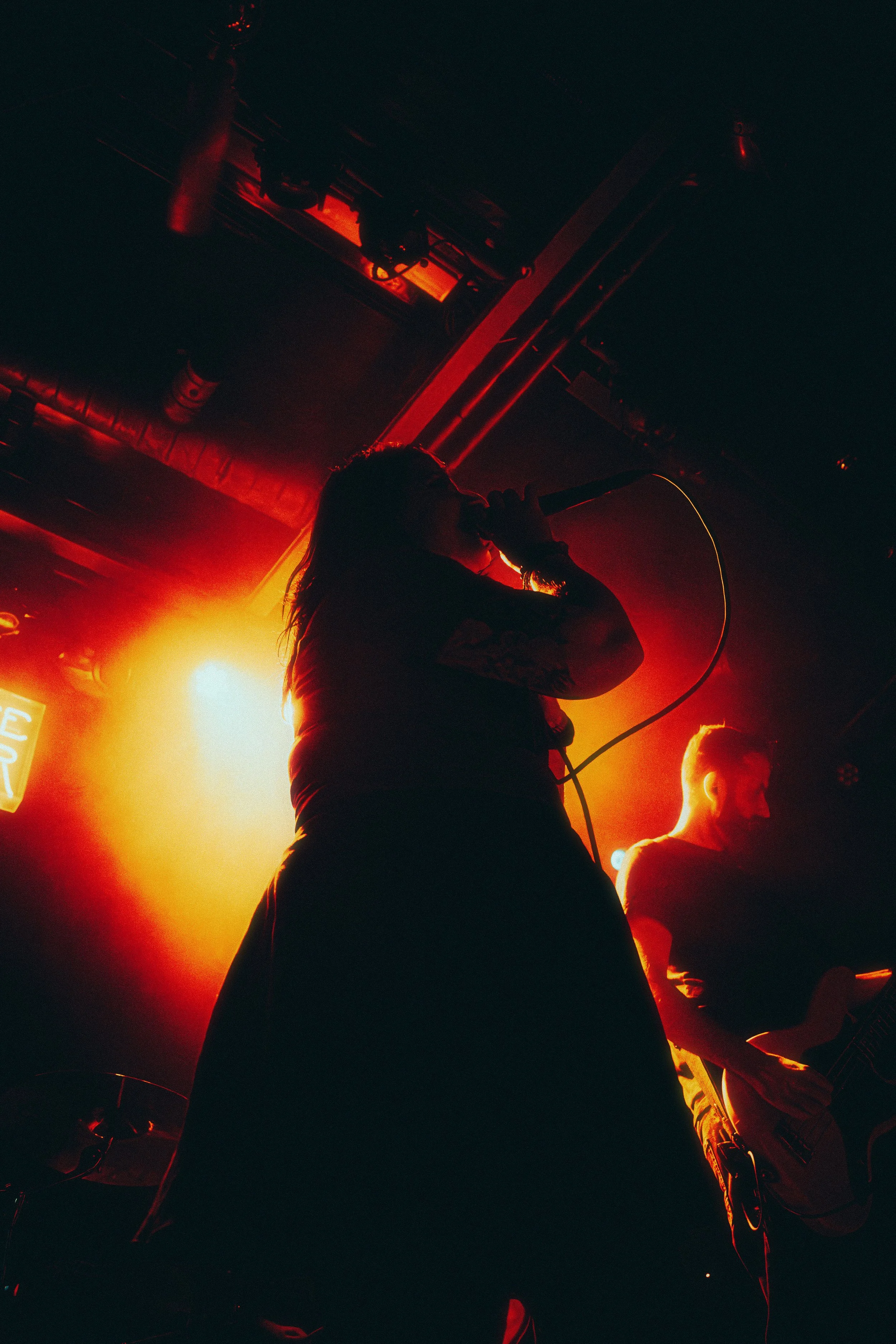 A silhouette of a vocalist with a microphone on stage illuminated by red and yellow stage lights, with a guitarist in the background.