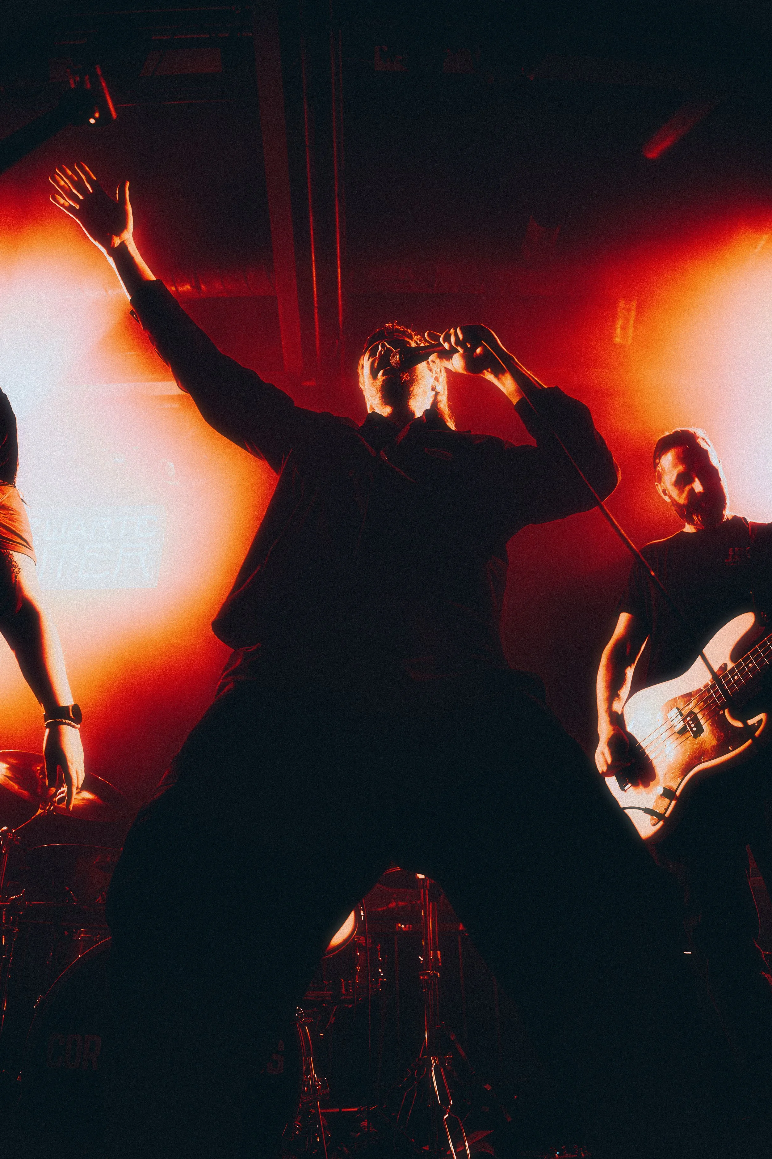 A vocalist singing into a microphone with a band performing on stage under red lighting.