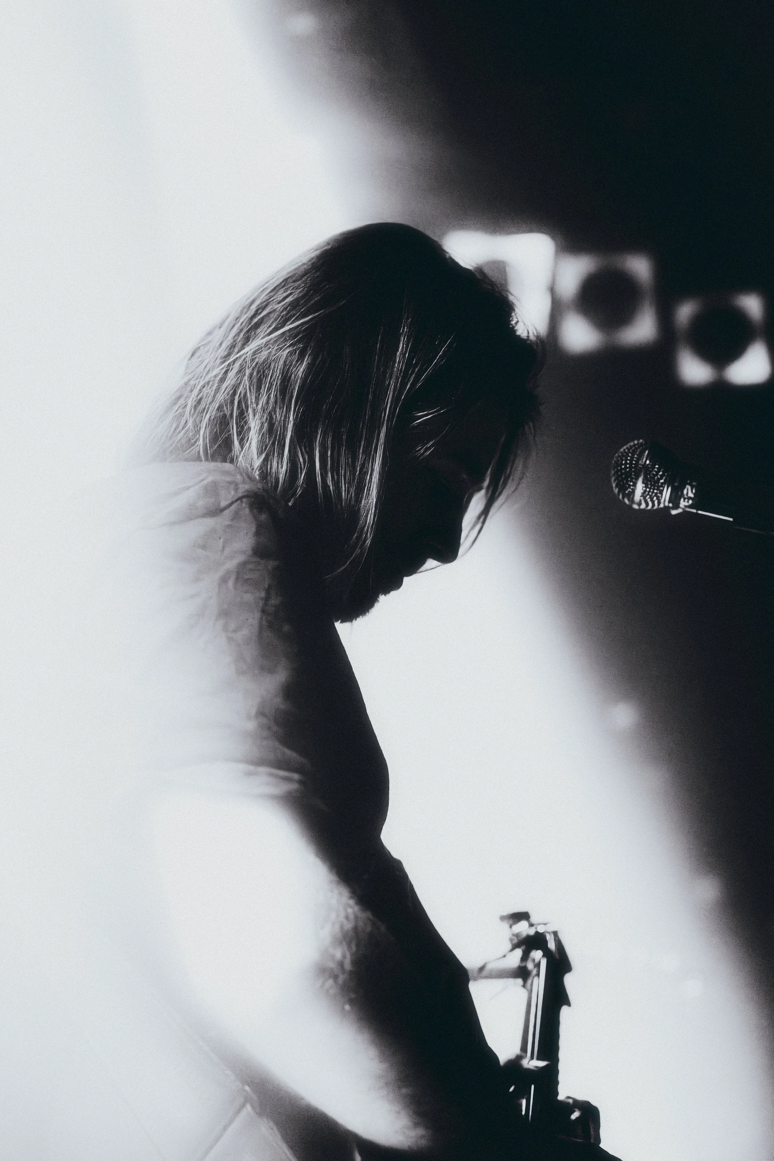 A black and white side profile of a person with long hair playing an electric guitar, with a microphone nearby and stage lights in the background.
