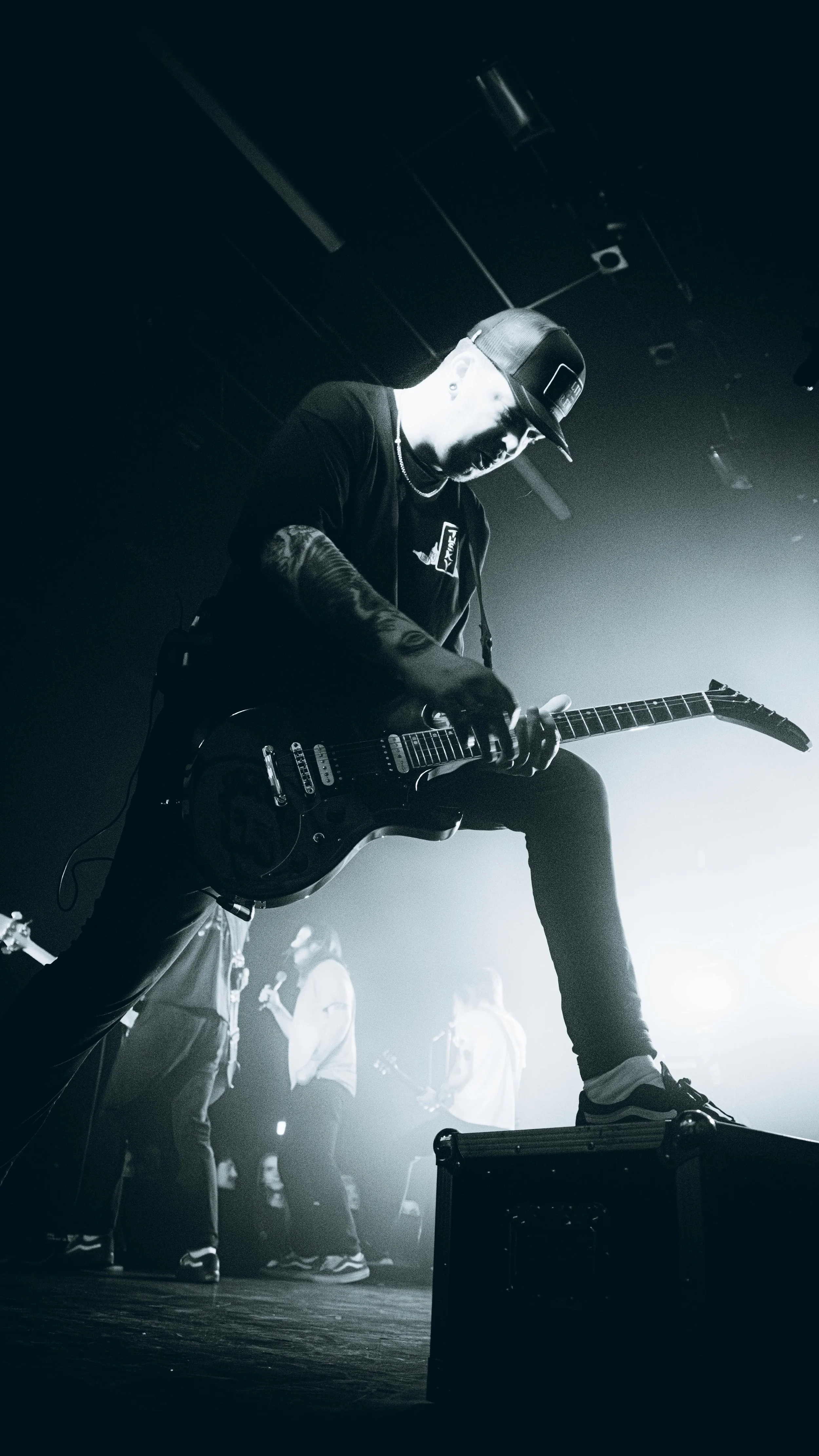 A musician playing an electric guitar on stage, with other band members in the background, in a black-and-white concert photo.