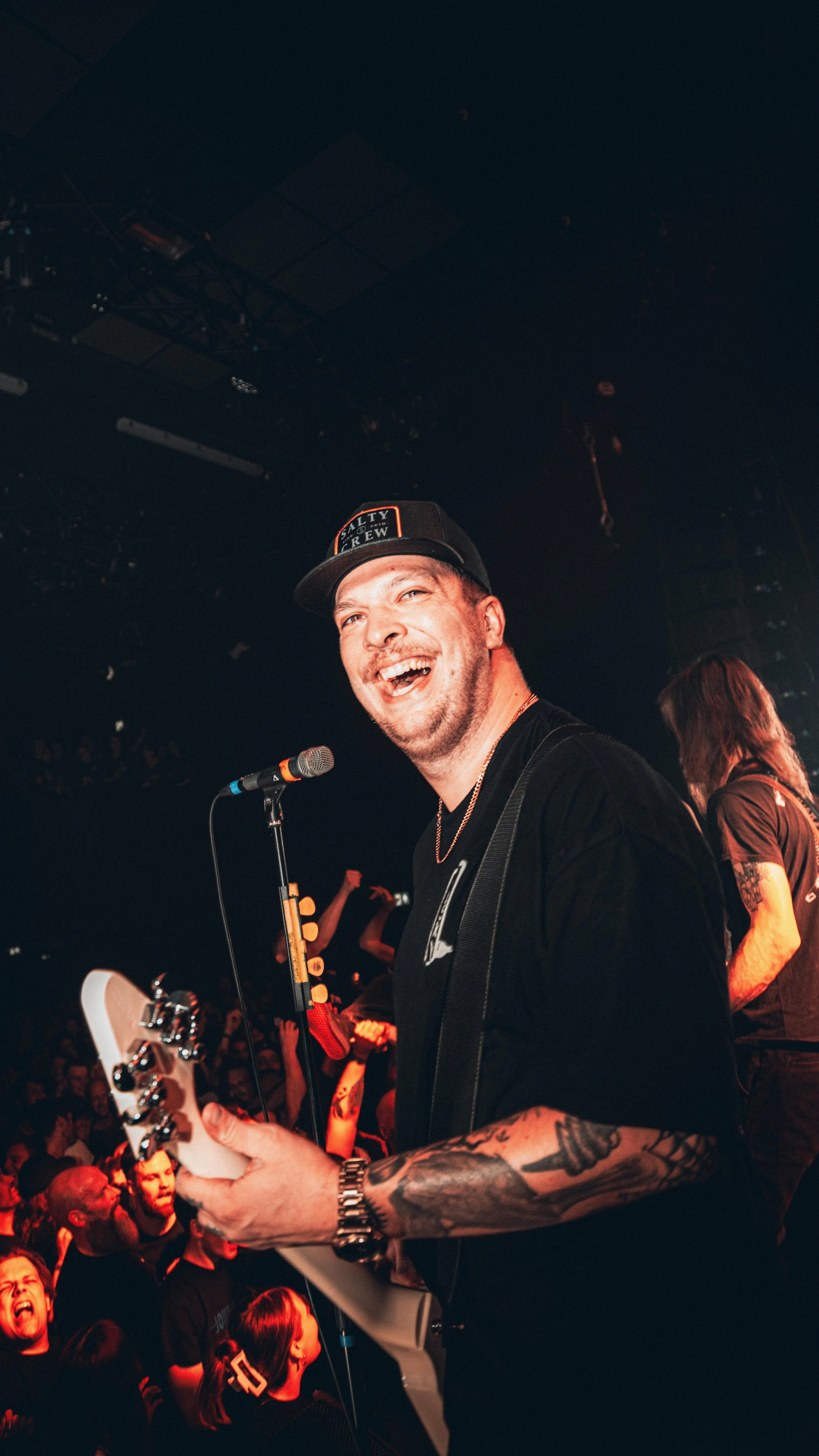 A man smiling and singing into a microphone while playing an electric guitar on stage at a concert, with a crowd in the background.