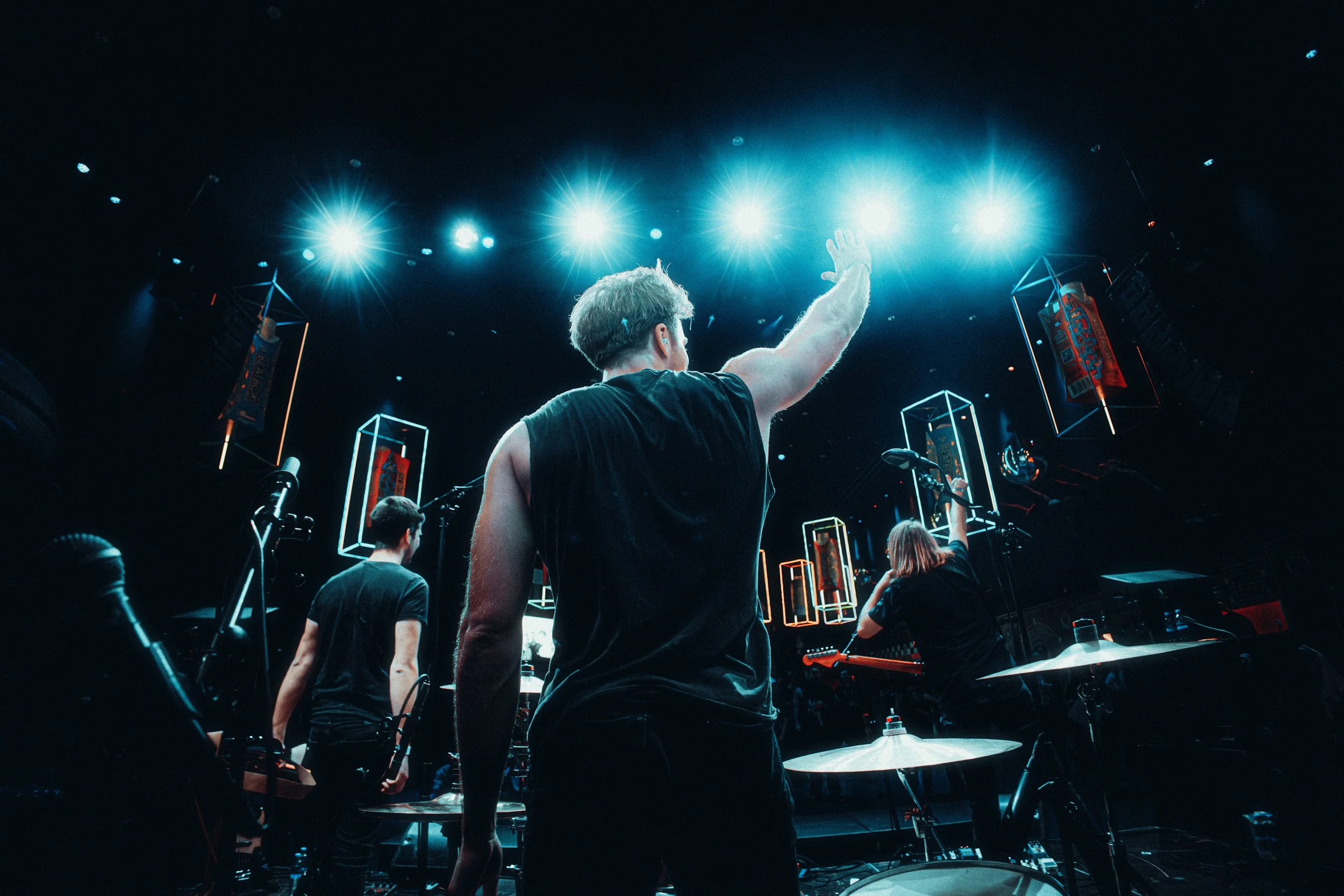 Musicians on stage during a concert with bright blue and white stage lights overhead, and decorative lighting boxes hanging in the background.