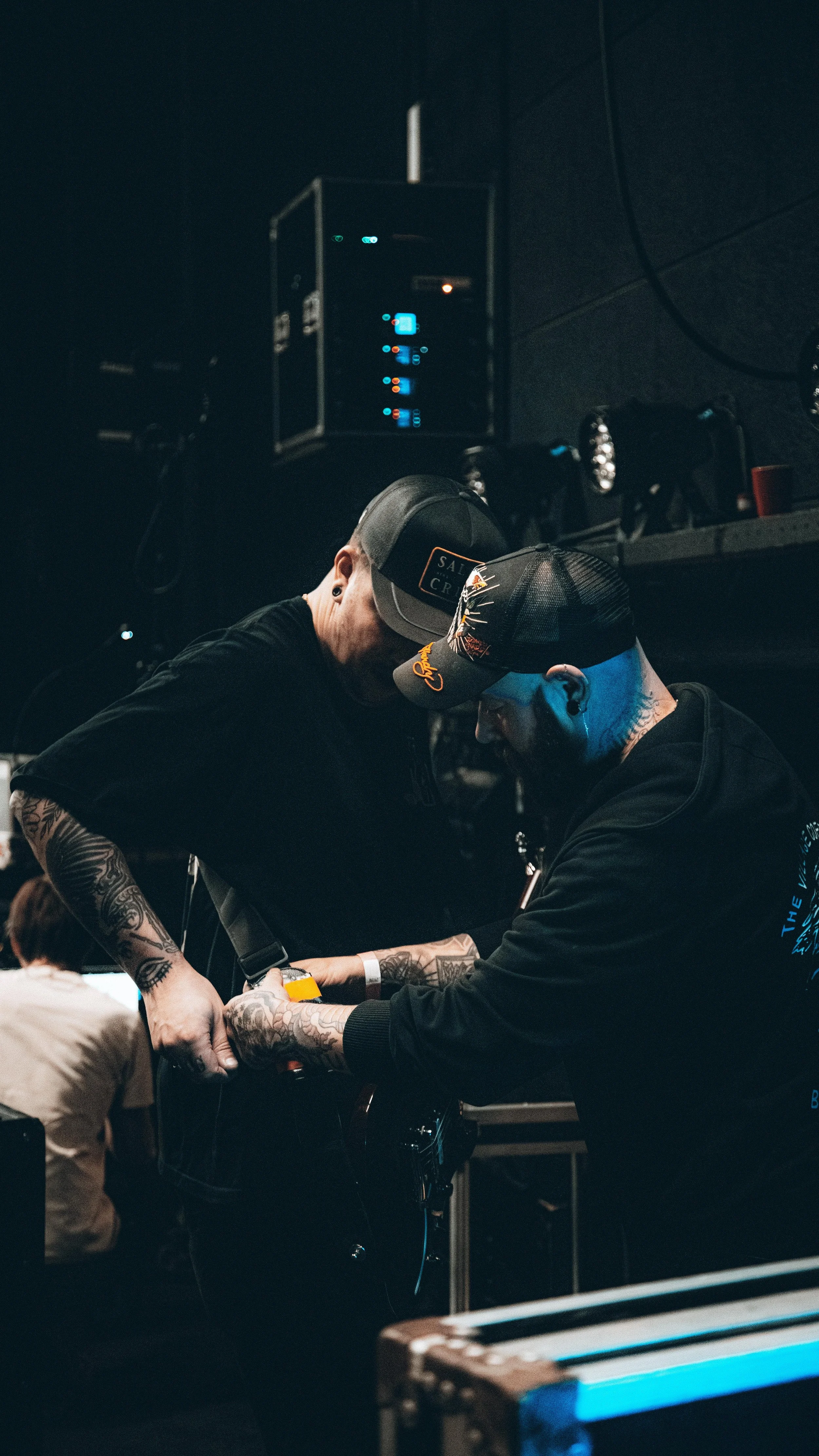 Two men with tattoos adjusting equipment in a dark room, likely a music or sound studio, with electronic equipment and lights in the background.