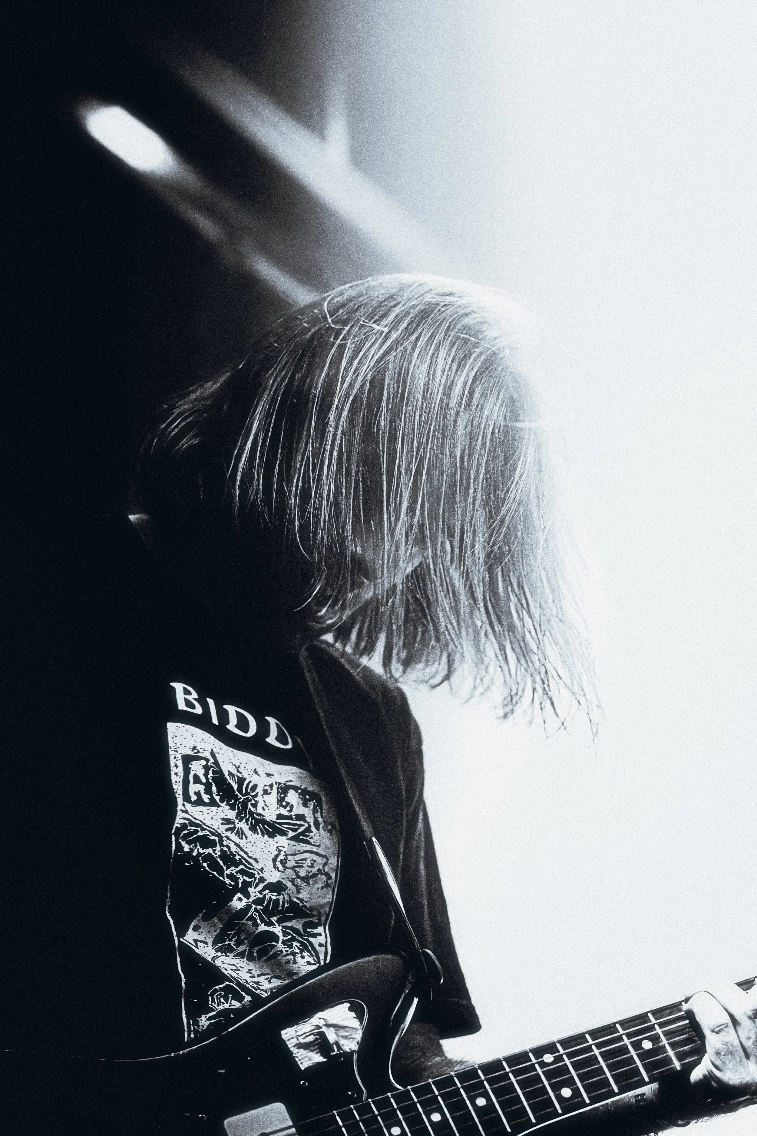 A person with long hair playing an electric guitar, wearing a black jacket with a graphic t-shirt that has the word "BID" on it, in black and white photography.