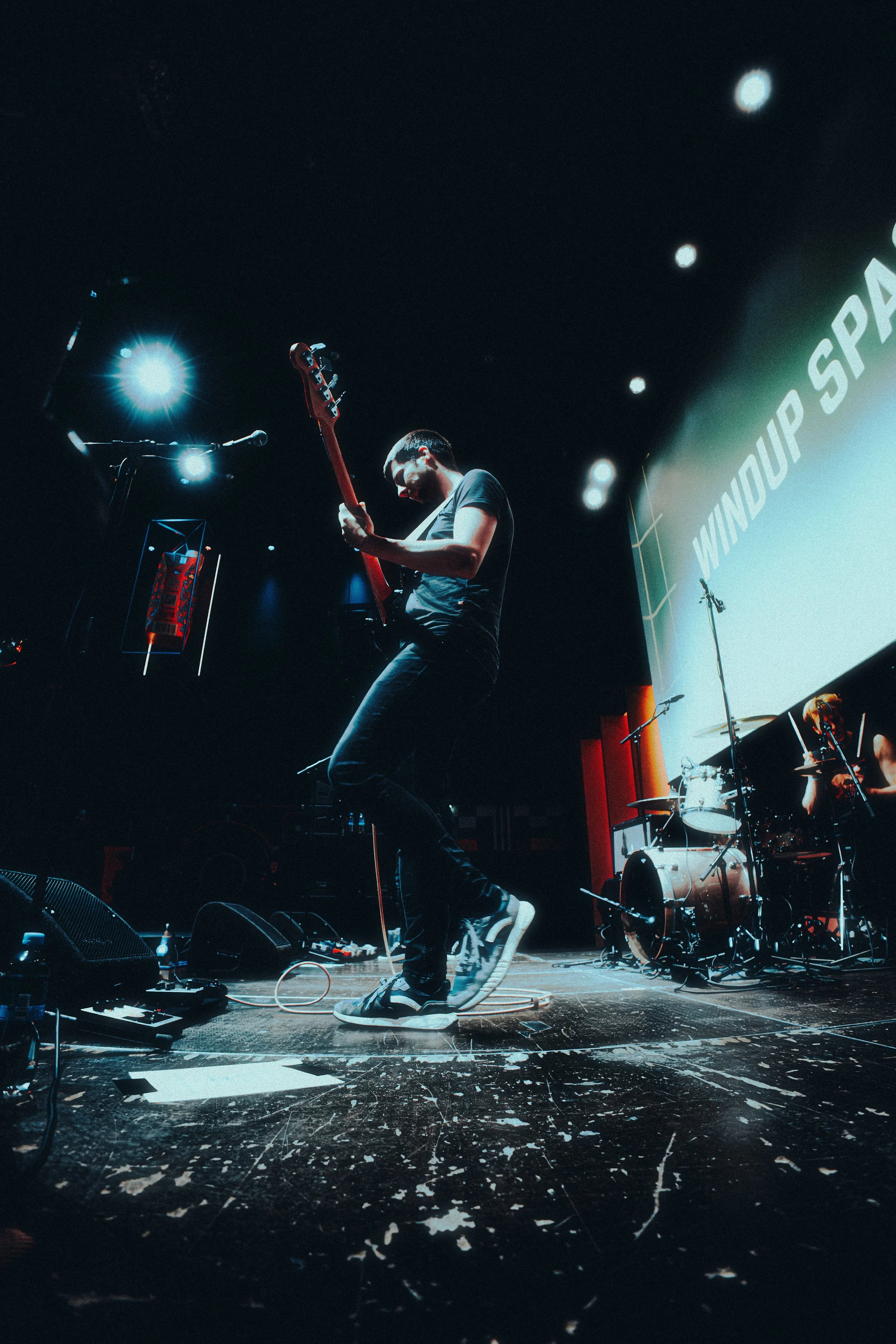 Musician playing electric guitar on stage during a performance, with stage lights and drums in the background at a Windows Up Space event.