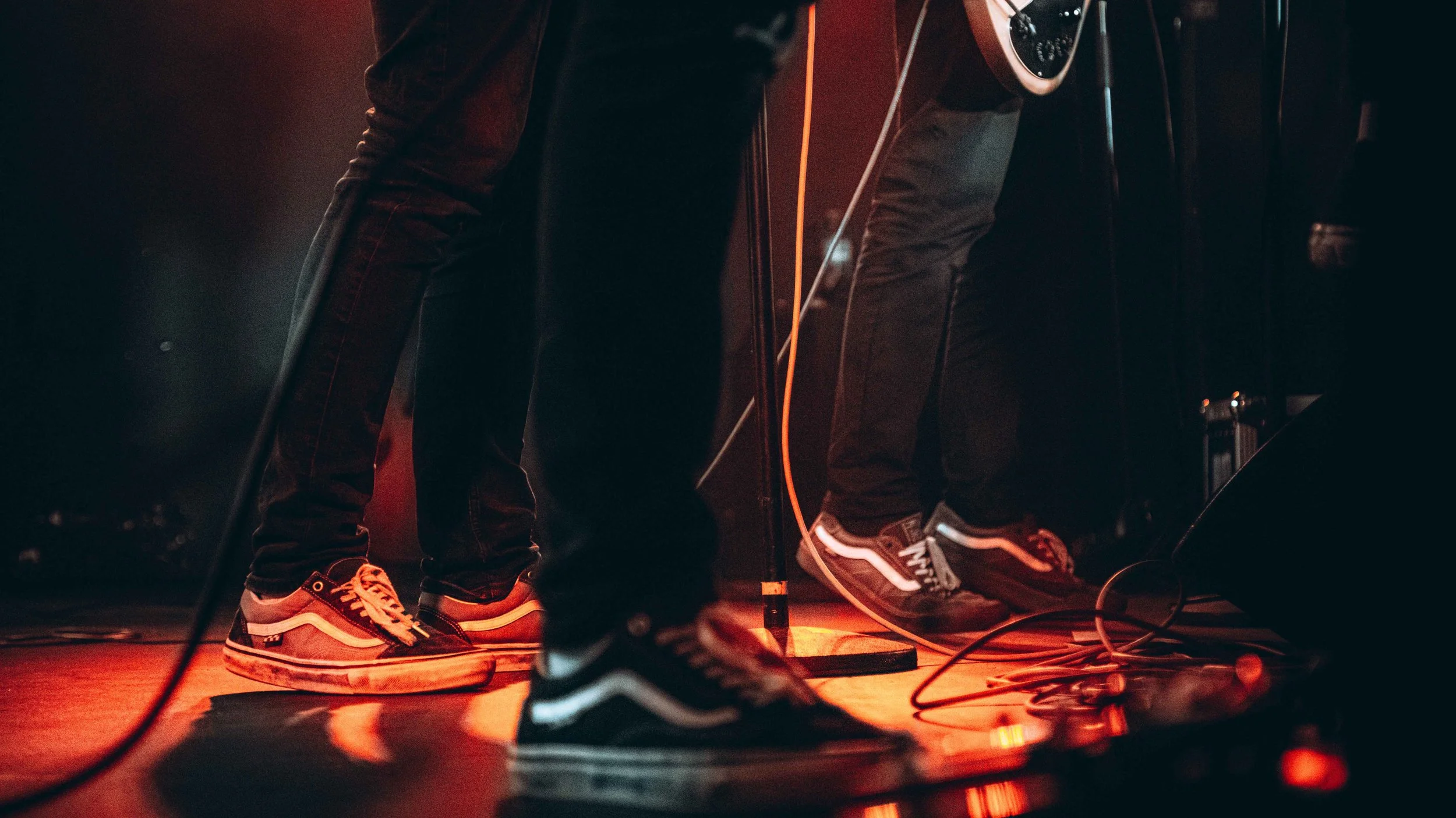 Close-up of two people's legs and feet wearing sneakers, performing on stage with guitars and microphone stand, illuminated by red stage lights.