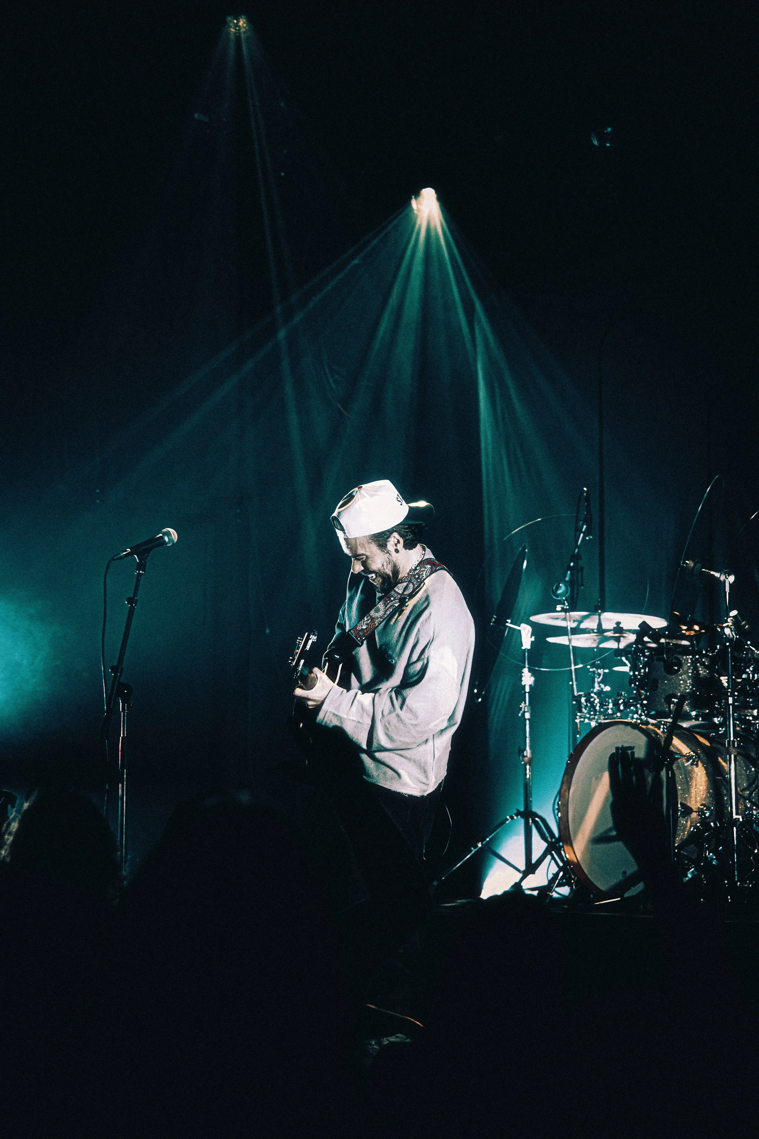 Musician playing guitar on stage with drum set in the background, illuminated by stage lights.