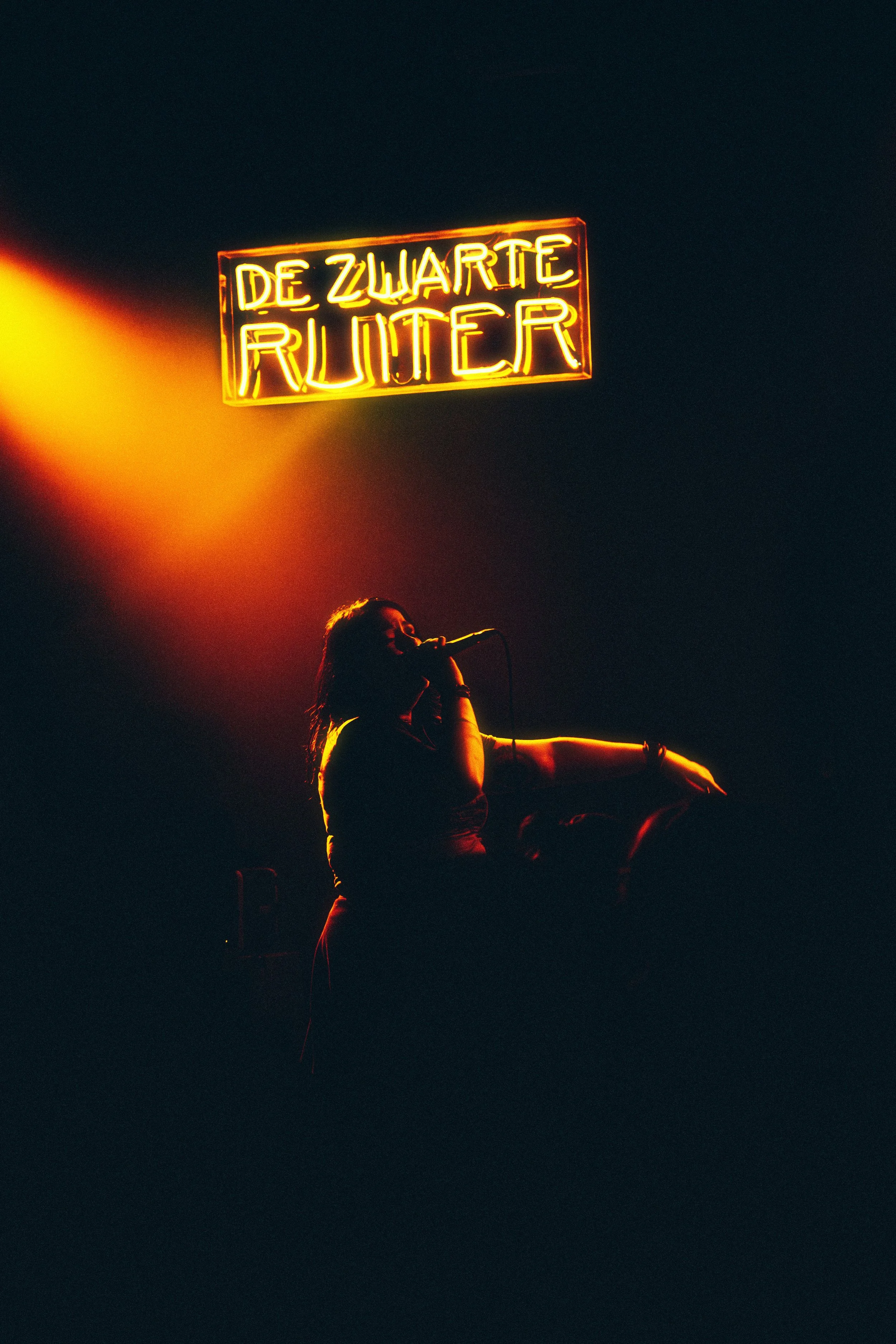 A woman singing into a microphone on stage with an orange spotlight, and a neon sign above her that reads 'De Zwarte Ruiter' in yellow and red neon lights.