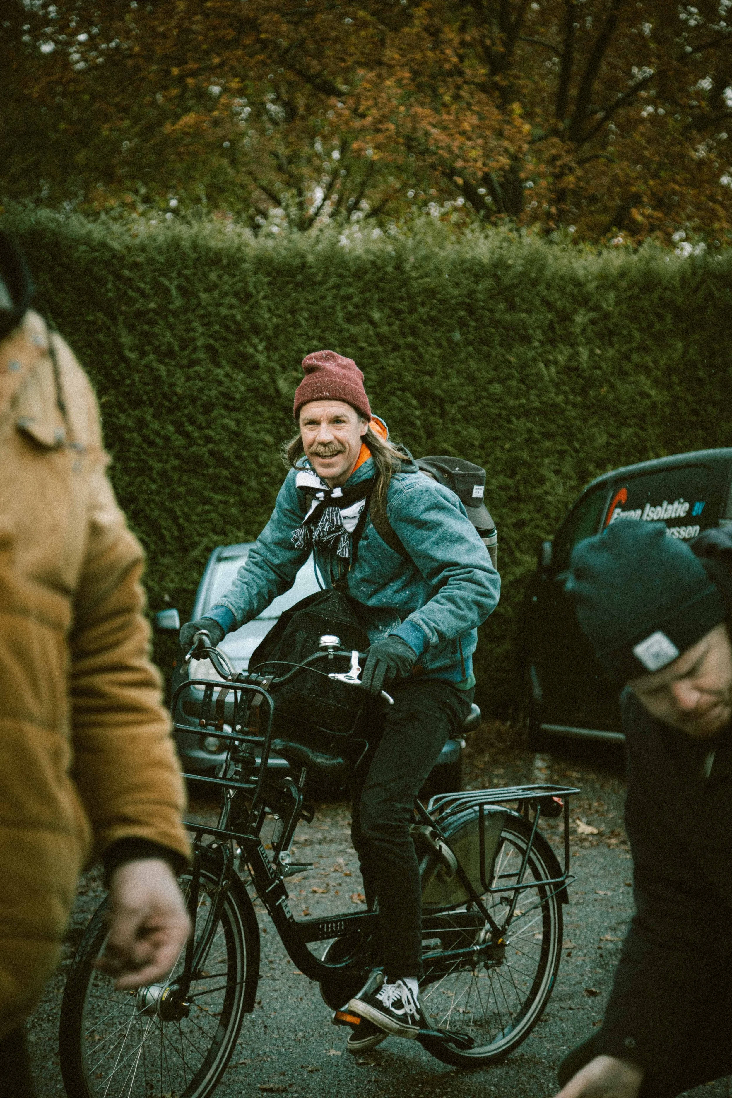 A man riding a bicycle outdoors, wearing a beanie, denim jacket, and gloves, surrounded by other people, with trees and parked cars in the background.