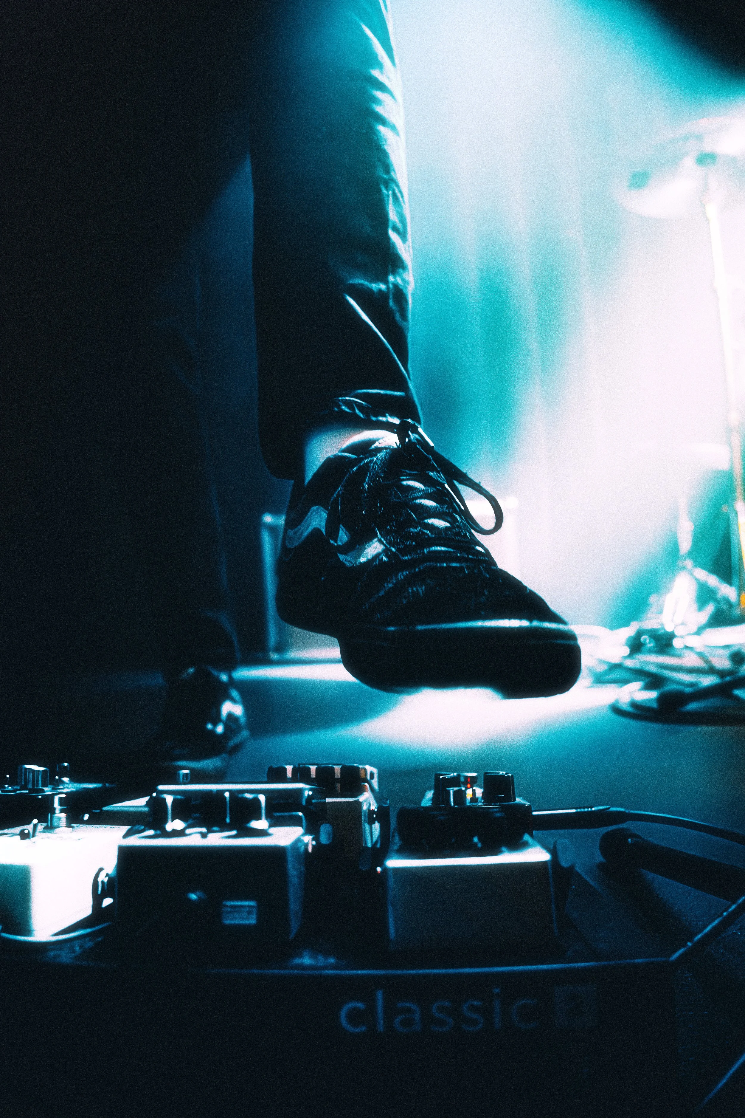 Close-up of a person's feet wearing shiny black shoes, standing on a DJ turntable with control knobs, illuminated by vibrant blue and white stage lighting.