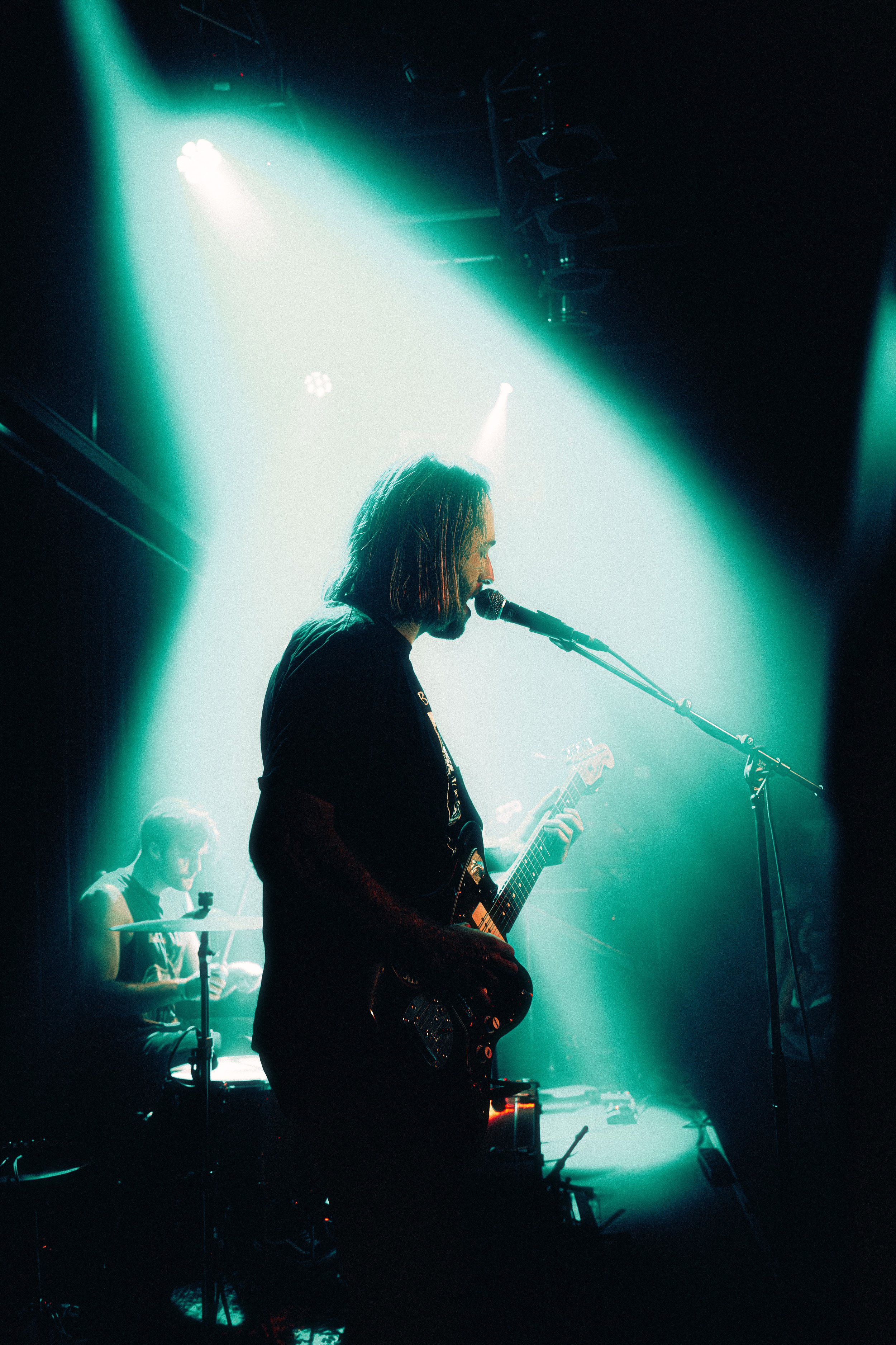 Musician with shoulder-length hair playing electric guitar and singing into a microphone, illuminated by stage lights, with a drummer in the background.