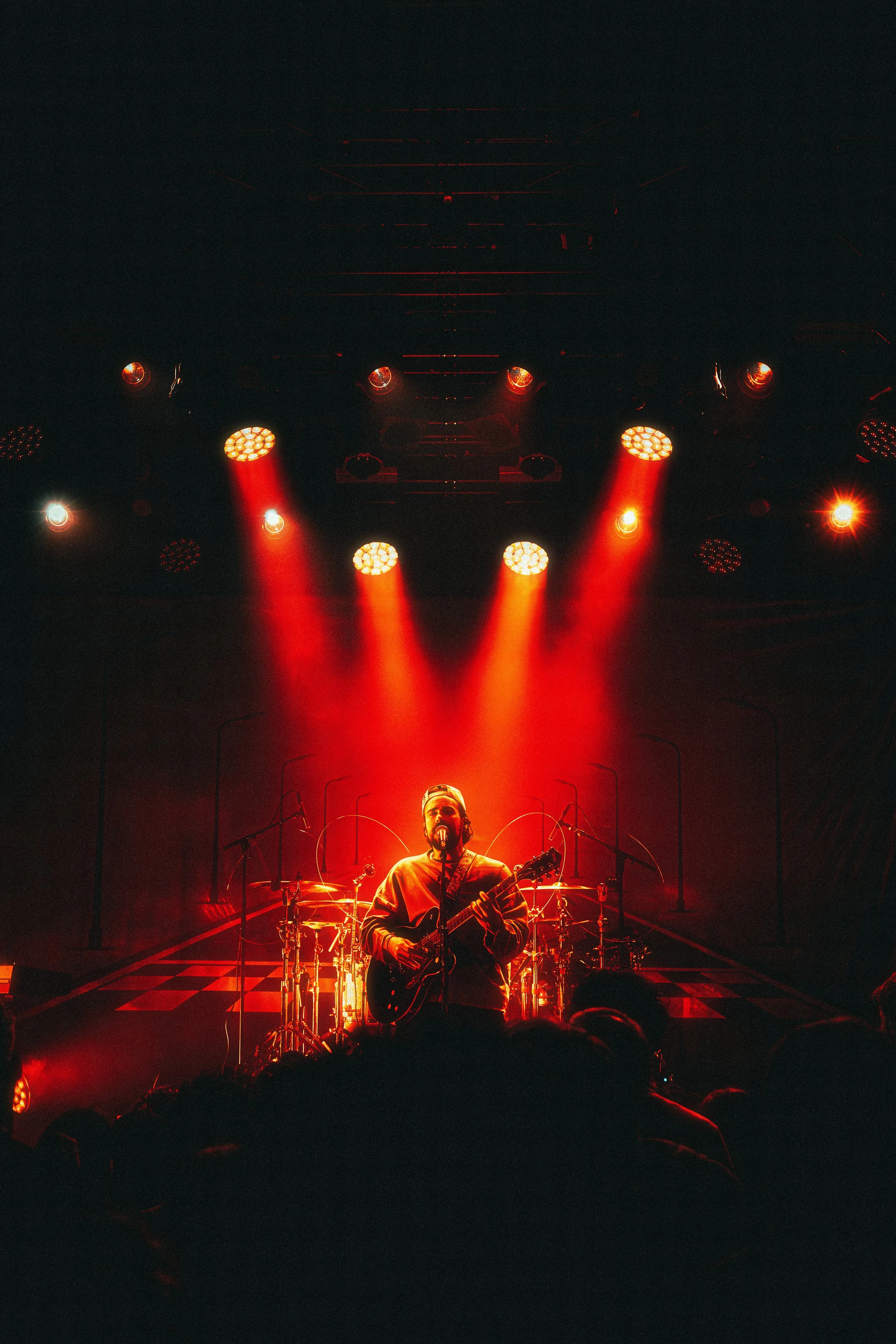 A musician performing on stage with a guitar, illuminated by red stage lights, with an audience in front.