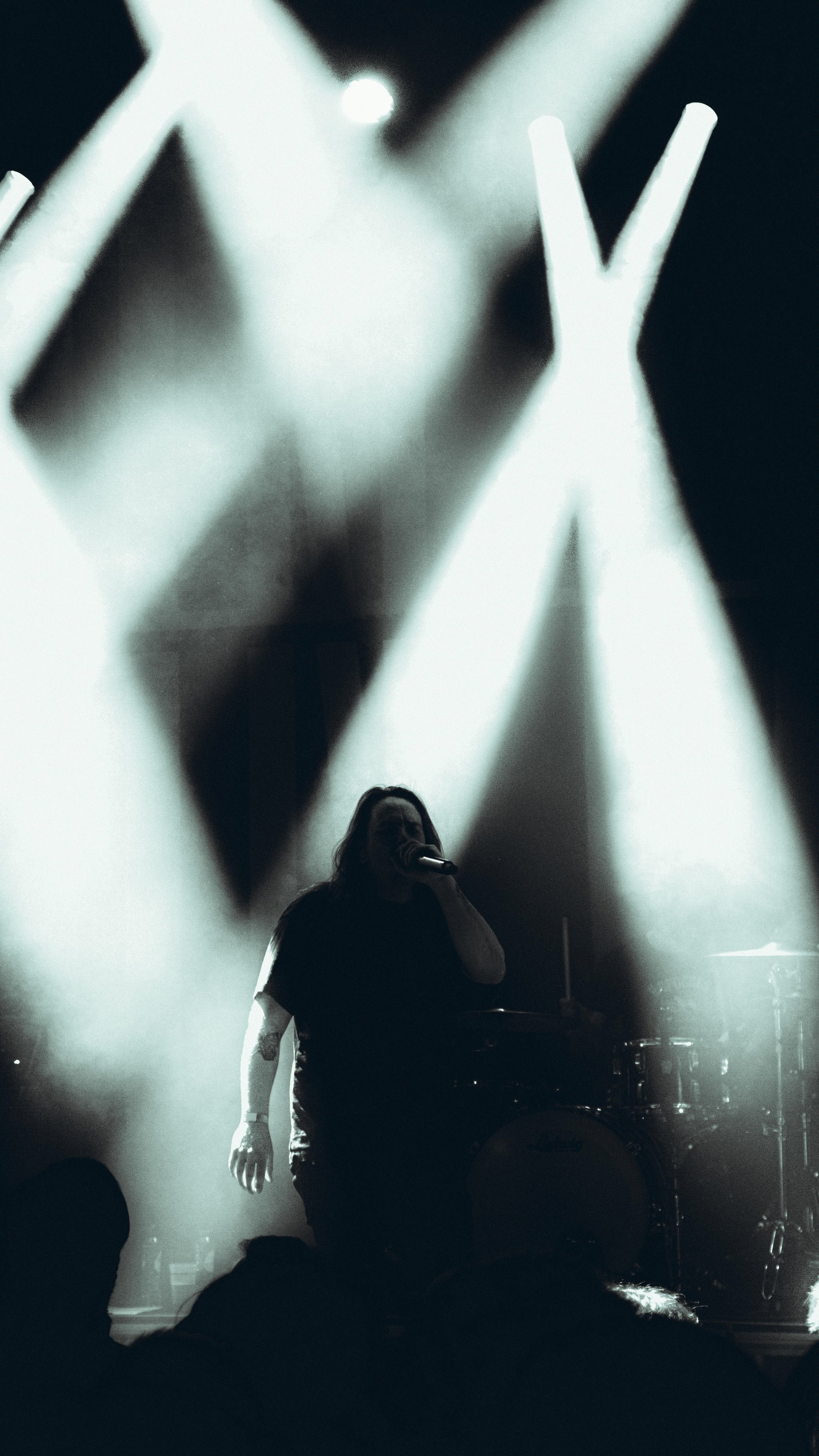 A person with long hair, holding a microphone, performing on stage with bright lights and a drum set in the background.