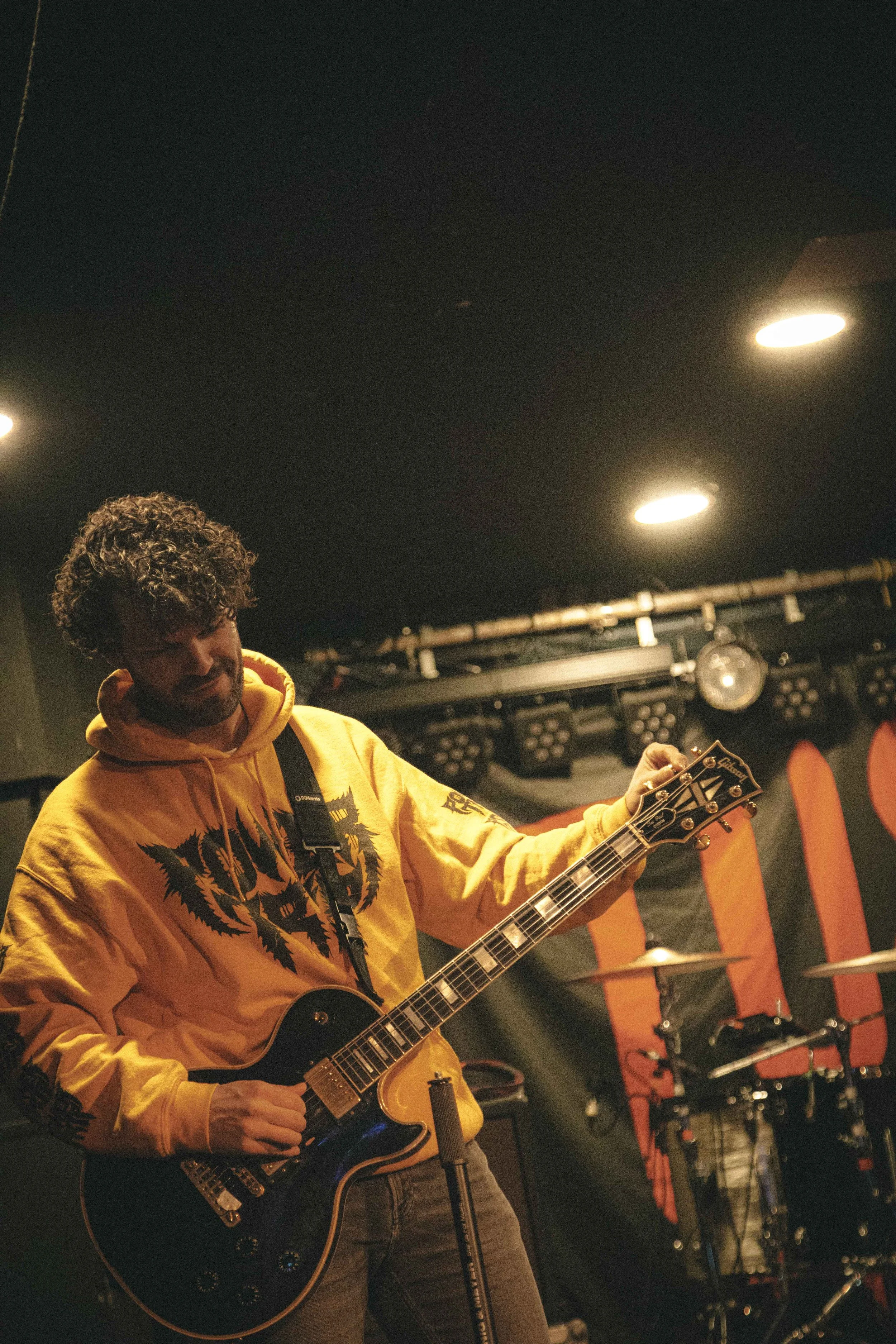 A man playing a black electric guitar on stage, wearing a yellow hoodie with a graphic design, in a dimly lit music venue.