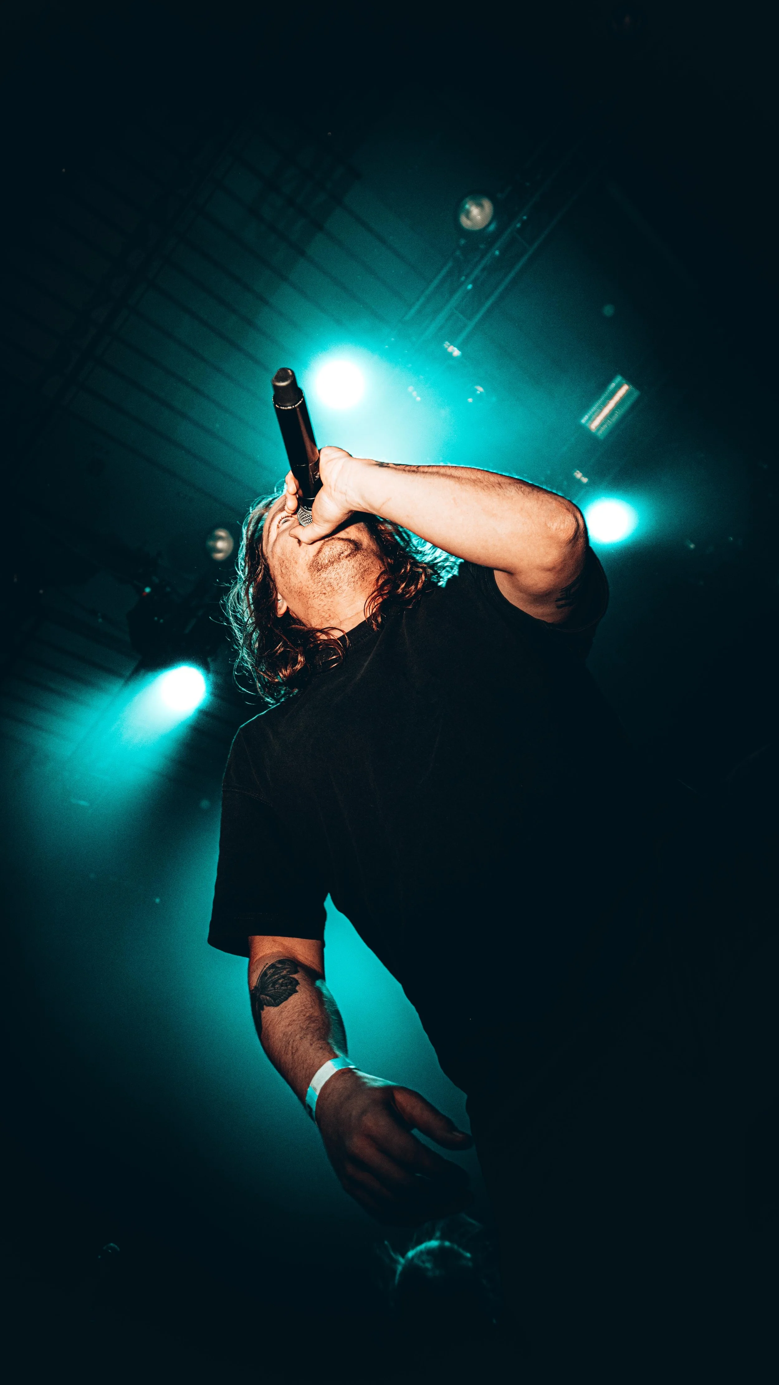 Male singer performing on stage with microphone, dark background with blue stage lights, tattoo on arm, wearing black t-shirt.