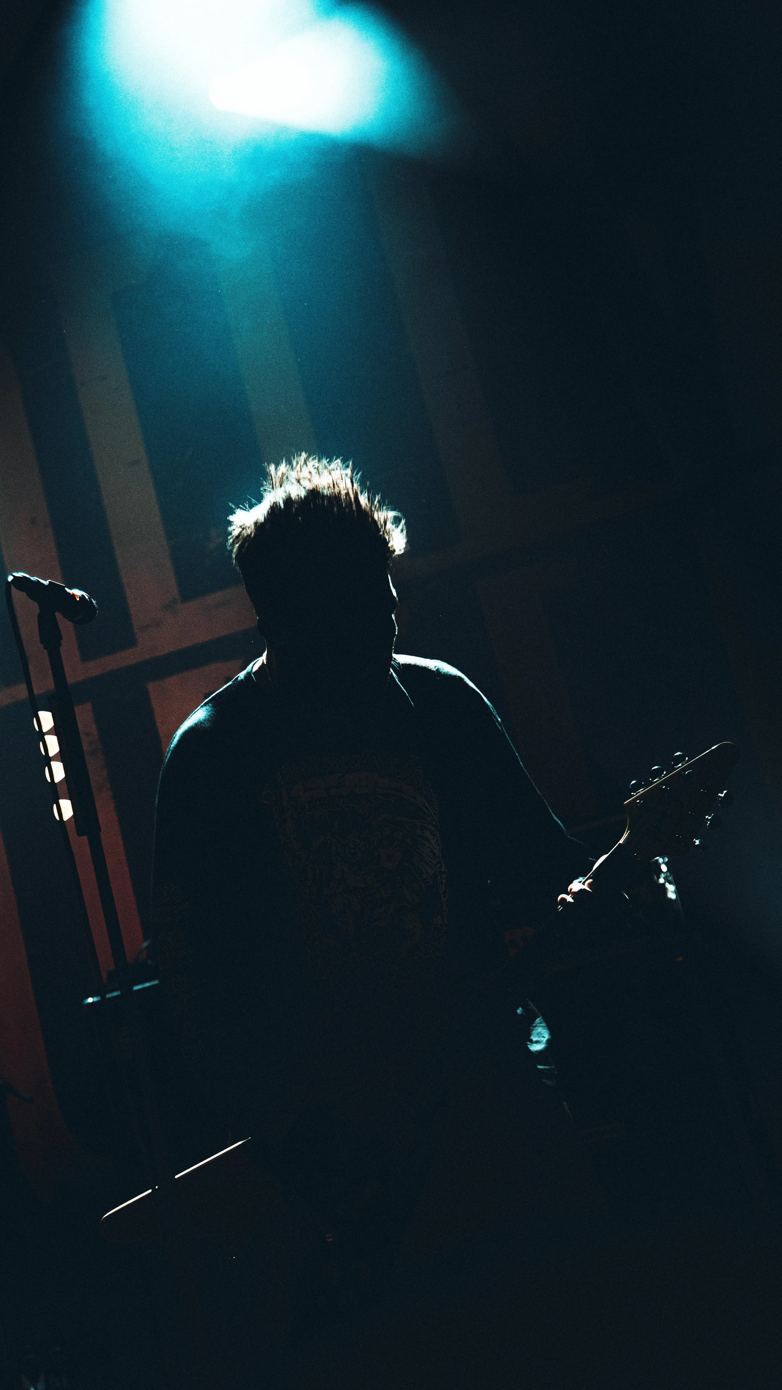 A silhouette of a person with spiky hair sitting in a dark room, illuminated by a bright blue light overhead, holding an instrument, possibly a guitar, with a microphone stand nearby.