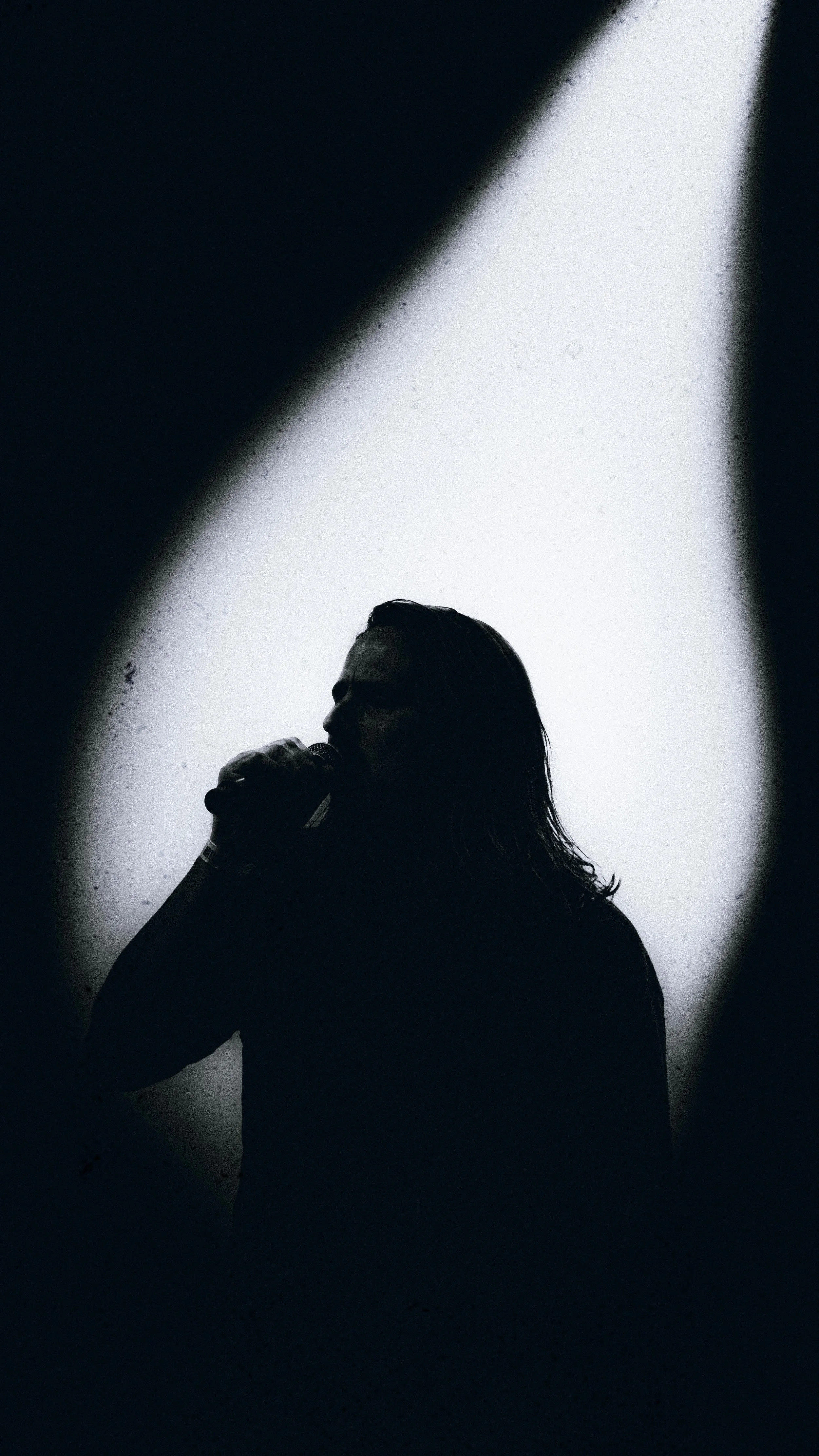 Silhouette of a person with long hair holding a microphone, seen through a dirty or foggy surface with light illuminating from behind.