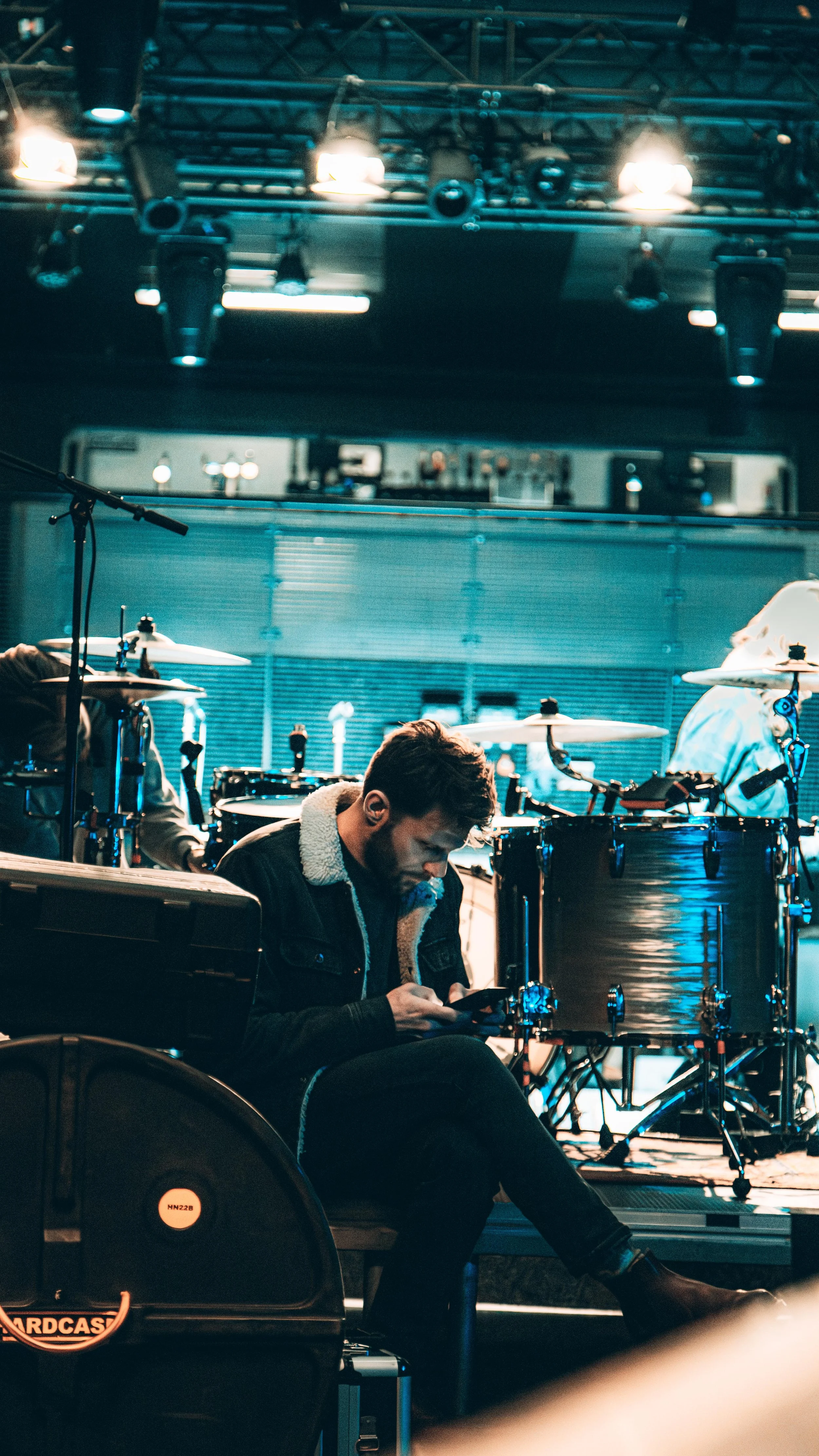 A person sitting on a bench in front of a drum set on a stage, looking at a smartphone, with stage lights overhead.