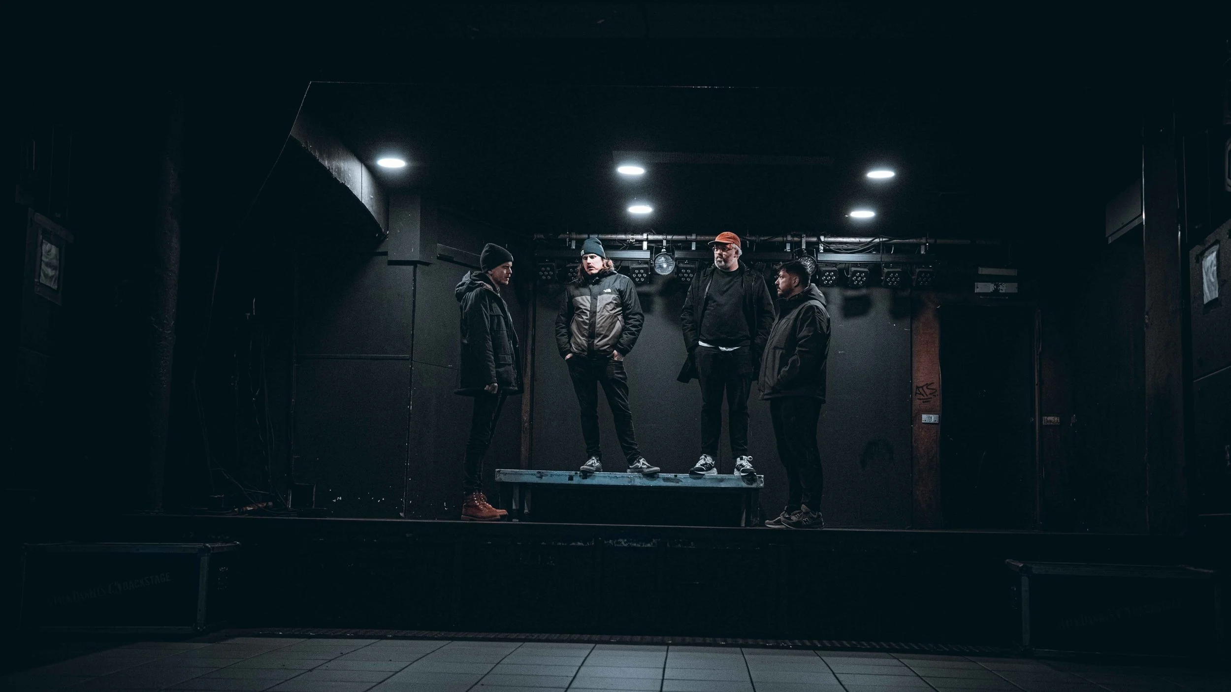 Four young men stand on a small elevated stage in a dark, empty venue, facing each other and dressed in casual winter clothing.