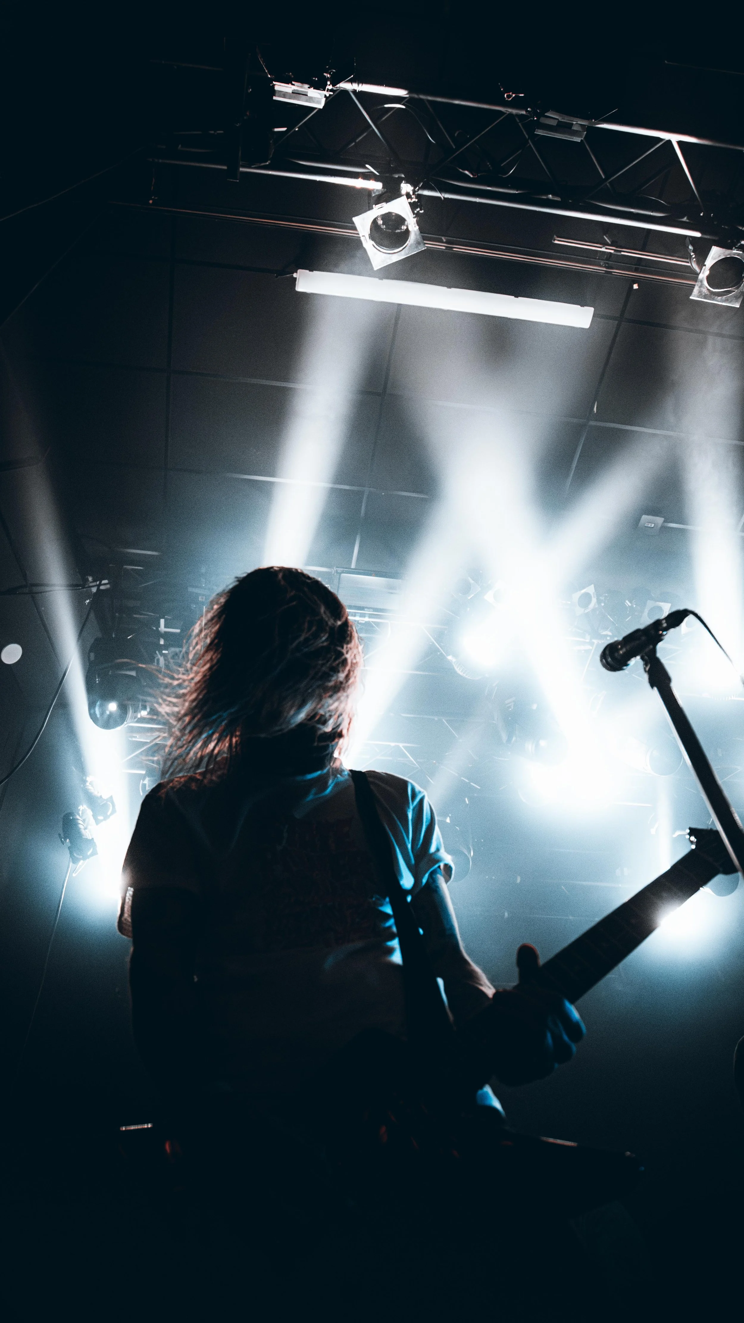 A musician playing an electric guitar on stage with bright stage lights shining behind them in a concert setting.