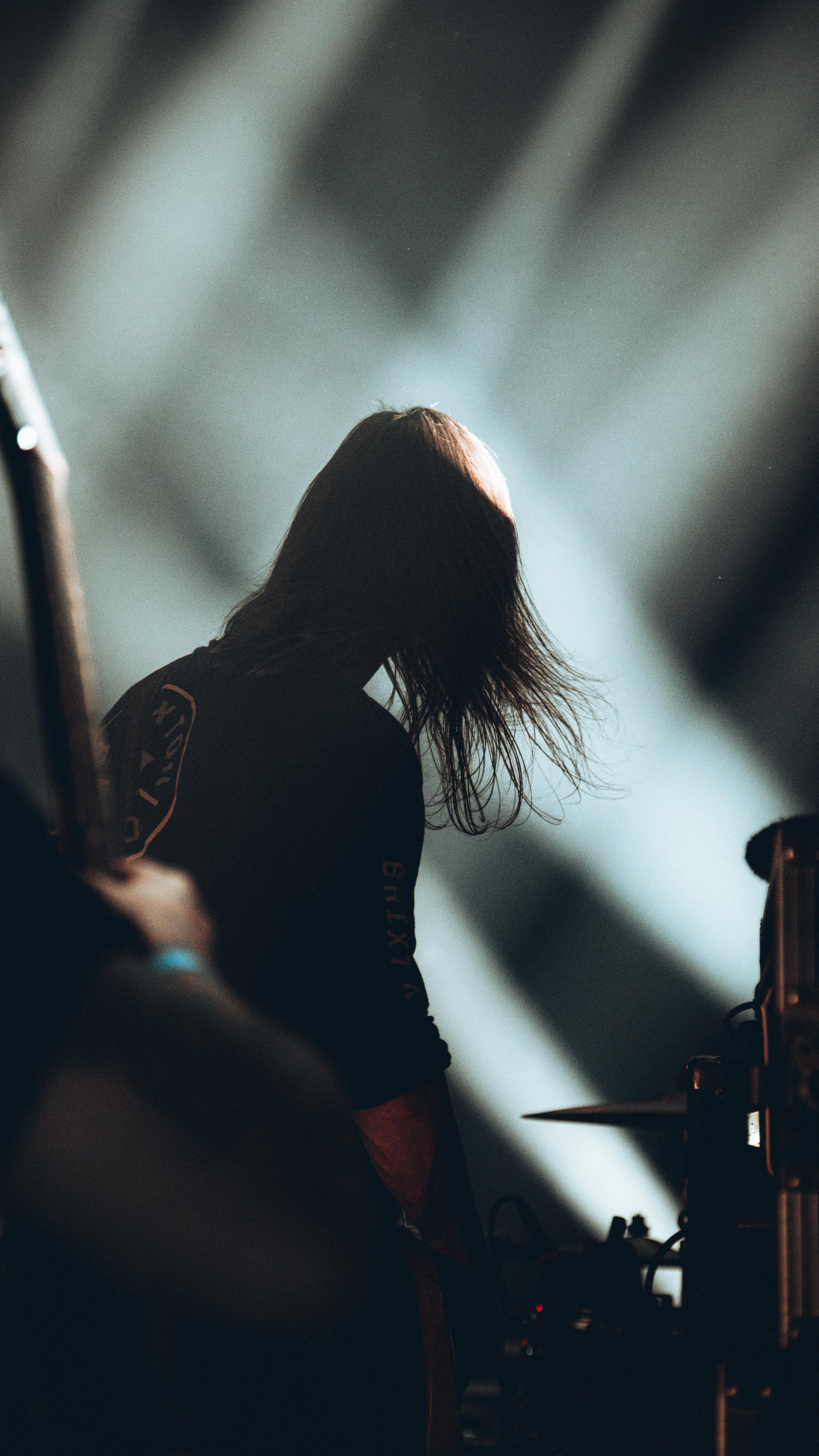 A person with long hair playing drums on a dimly lit stage, with light shining from behind.