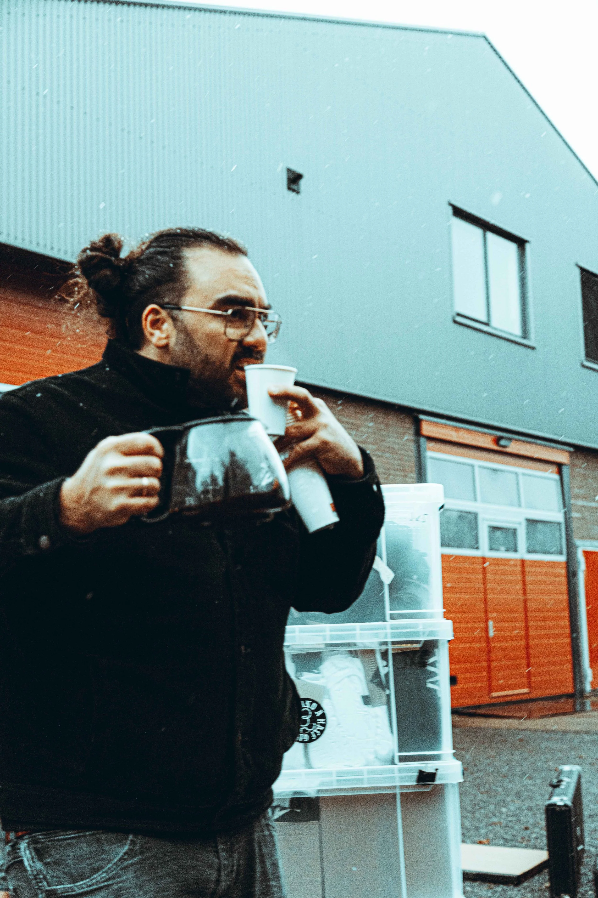 A man with glasses and a man bun drinking from a cup while holding a coffee pot, standing outdoors near a building with orange and gray siding.