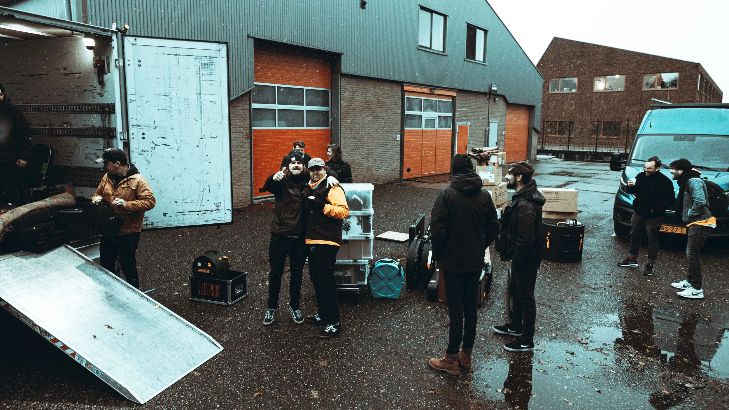Group of people loading and unloading a vehicle in an outdoor area with wet pavement, brick and metal buildings, and a parked van.
