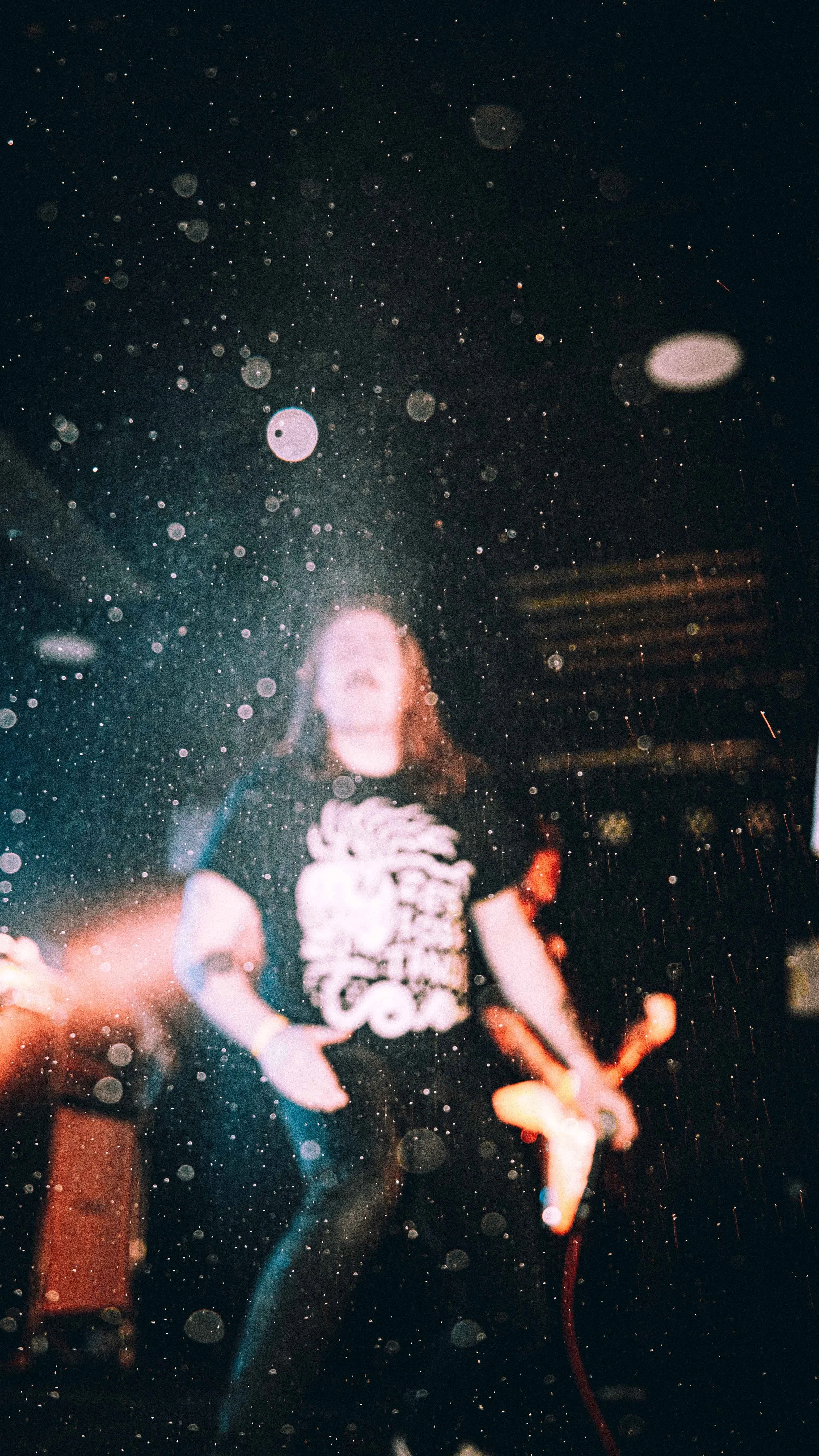 A woman standing behind a rain-covered window at night, with city lights and blurred buildings in the background.