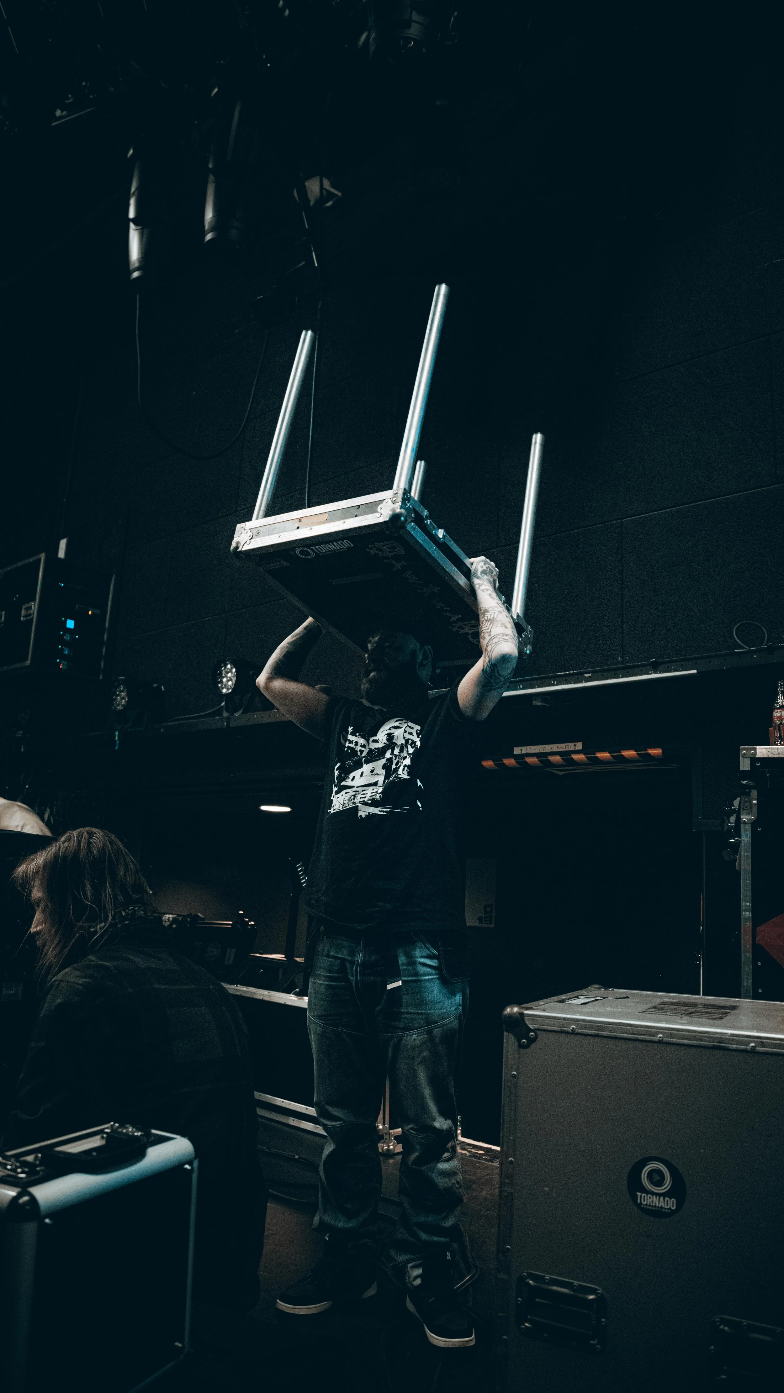 A person wearing a black t-shirt and jeans lifting a table or platform with poles on a stage or studio, with equipment and cables around.
