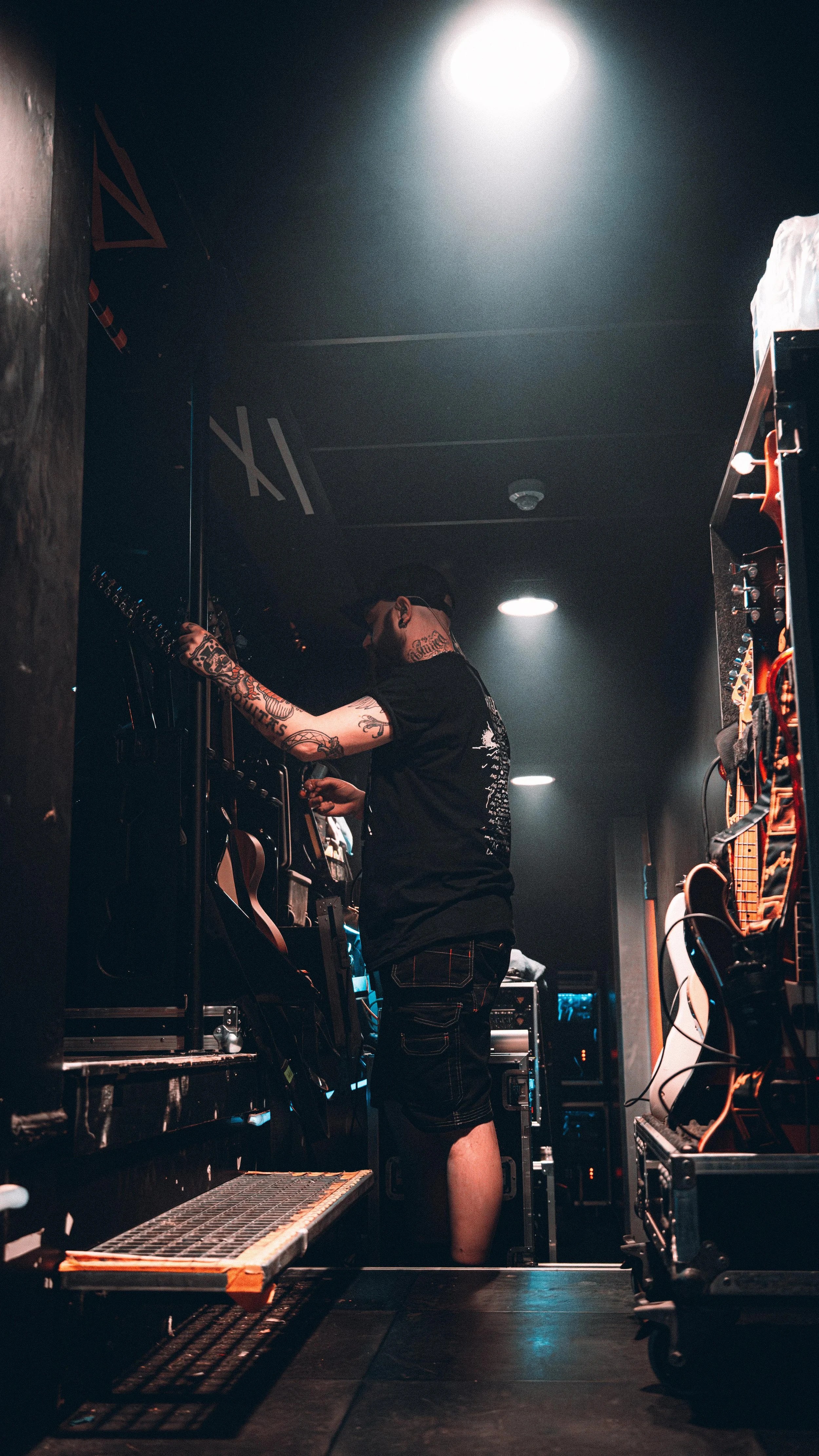 A man with tattoos on his arms and neck adjusting equipment backstage at a music venue.