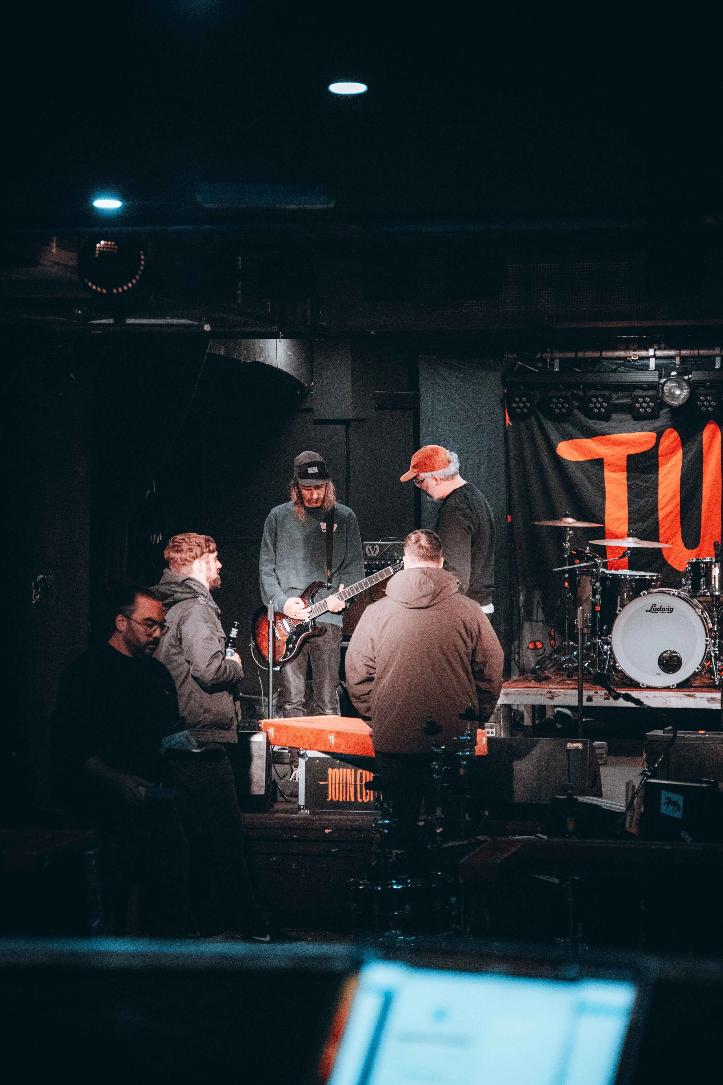 A band practicing on stage in a dimly lit venue, with four men near musical instruments including a keyboard, guitar, and drums.
