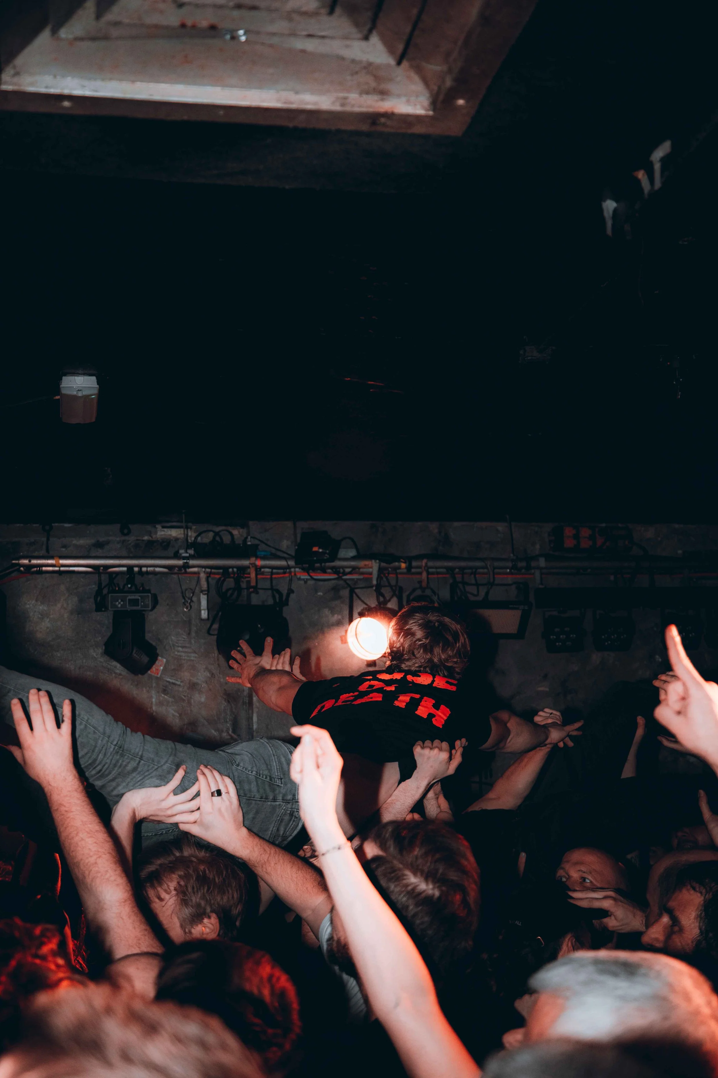 Crowd surfing at a concert, person in black T-shirt with red lettering being held up by hands, stage lighting, and audience members reaching out.