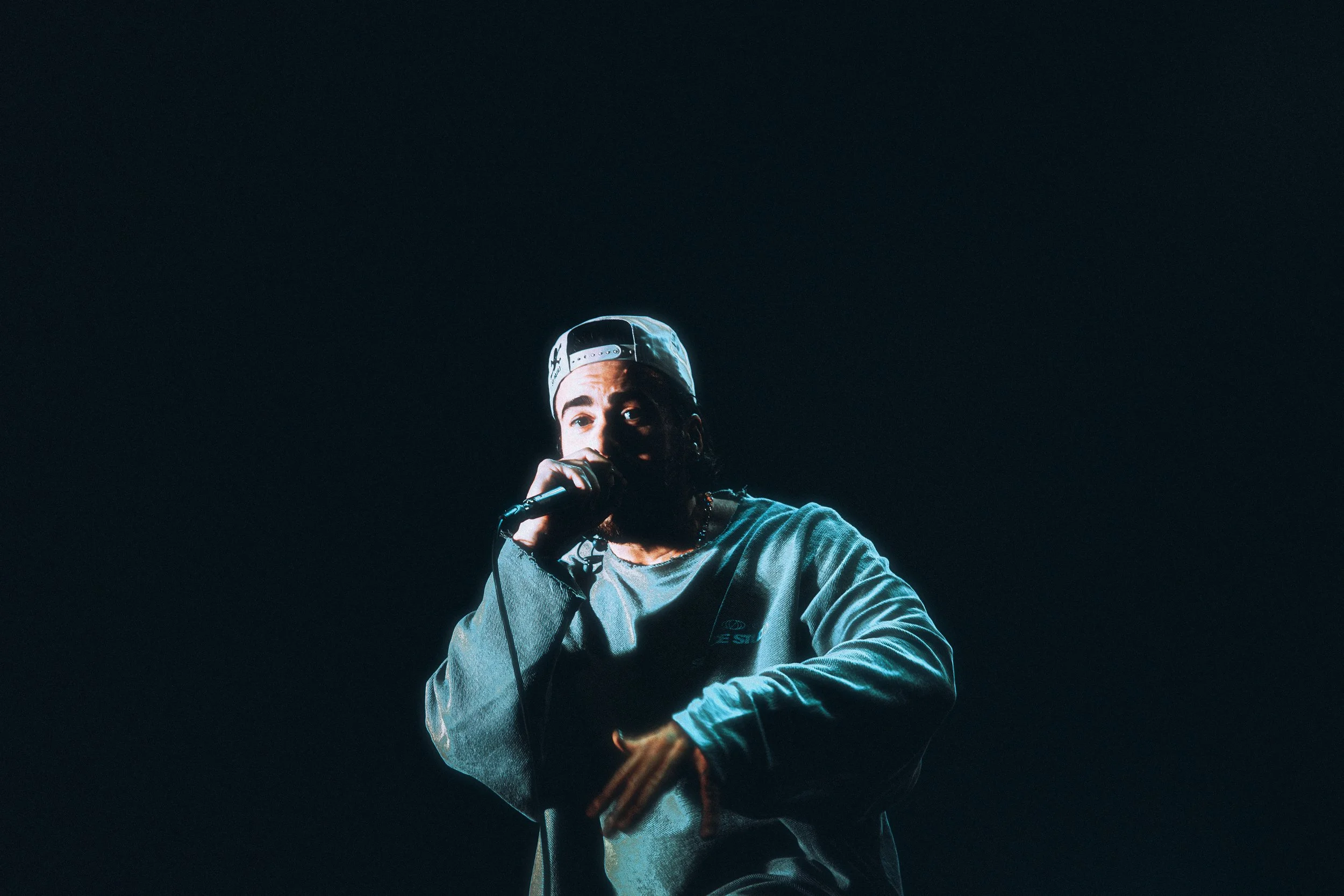 Young man performing with a microphone on stage, wearing a backward cap and casual clothing, illuminated by stage lighting against a dark background.