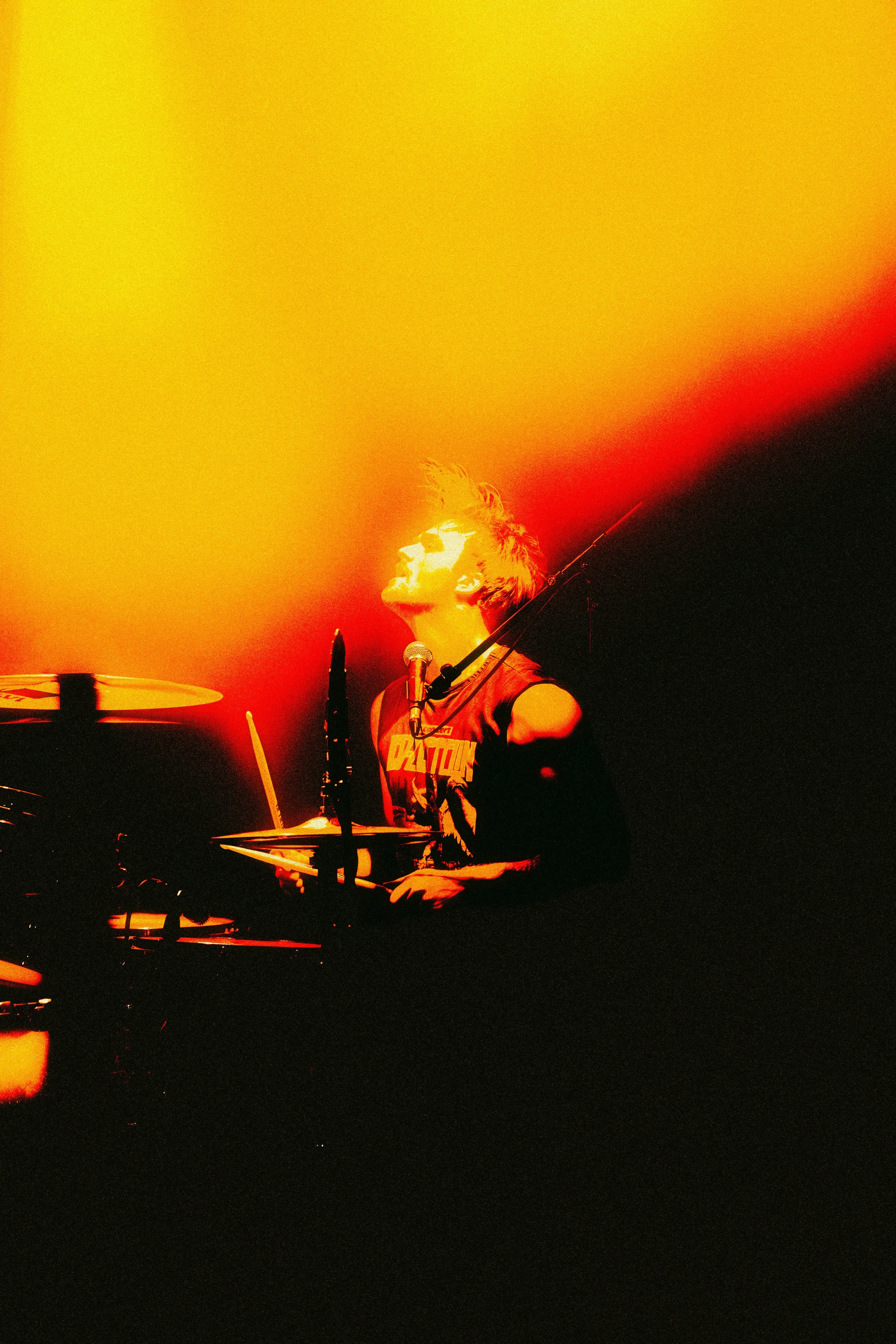 A drummer performing on stage, illuminated by red, orange, and yellow lights, with a focused expression.