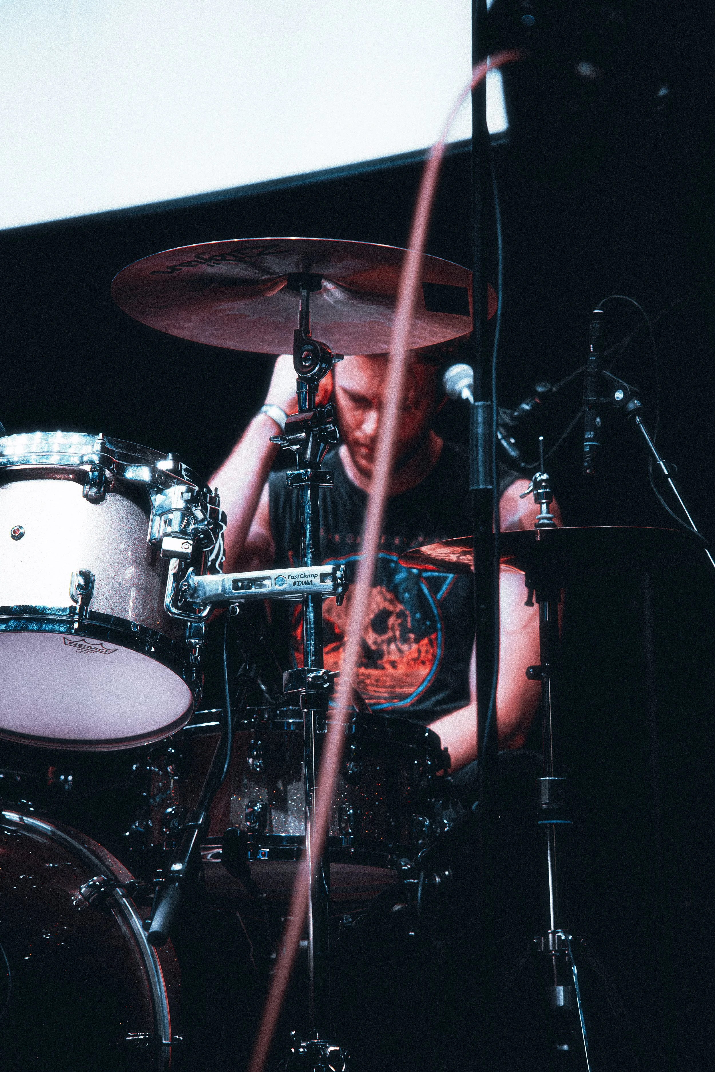 A drummer playing a drum set on stage, with a focused expression, wearing a black sleeveless shirt, and surrounded by microphones and stage equipment.