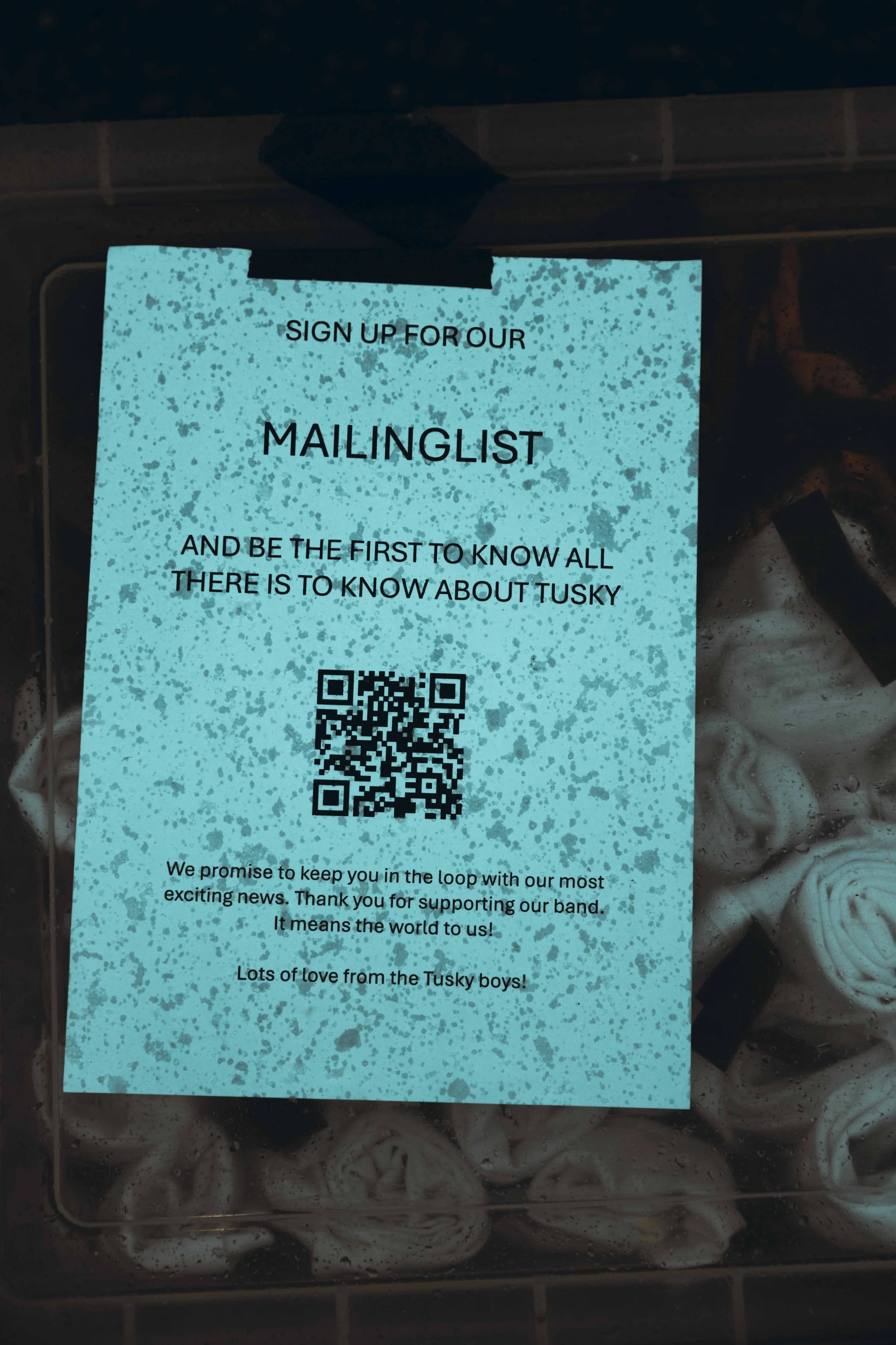 A blue sign with black speckles requesting people to sign up for a mailing list, featuring a QR code and messages about supporting a band called the Tusky boys, inside a plastic container filled with marshmallows.