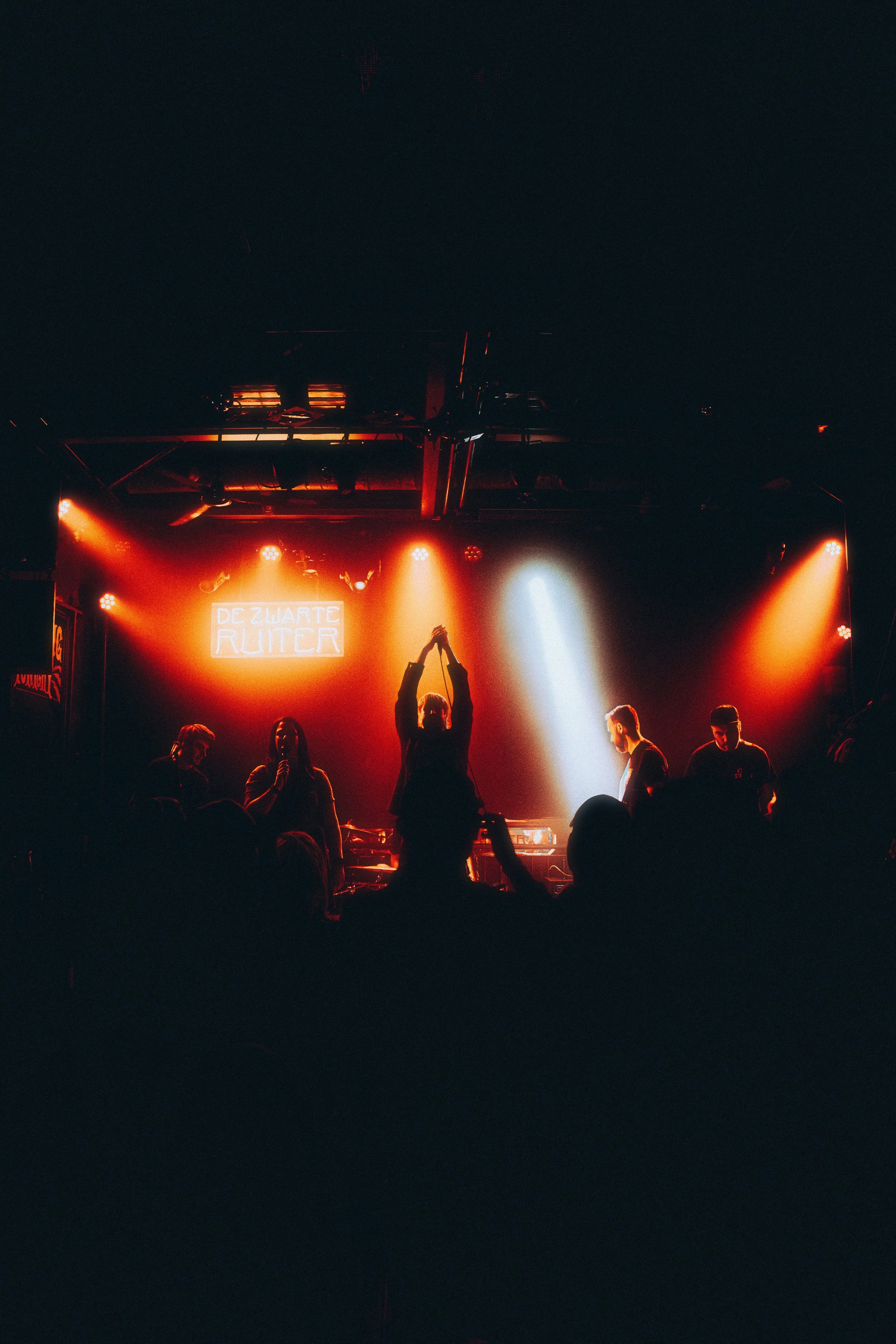 People performing on stage with red and white lights at a live music event, with a sign reading "Dezwarte Ruiter" in the background.