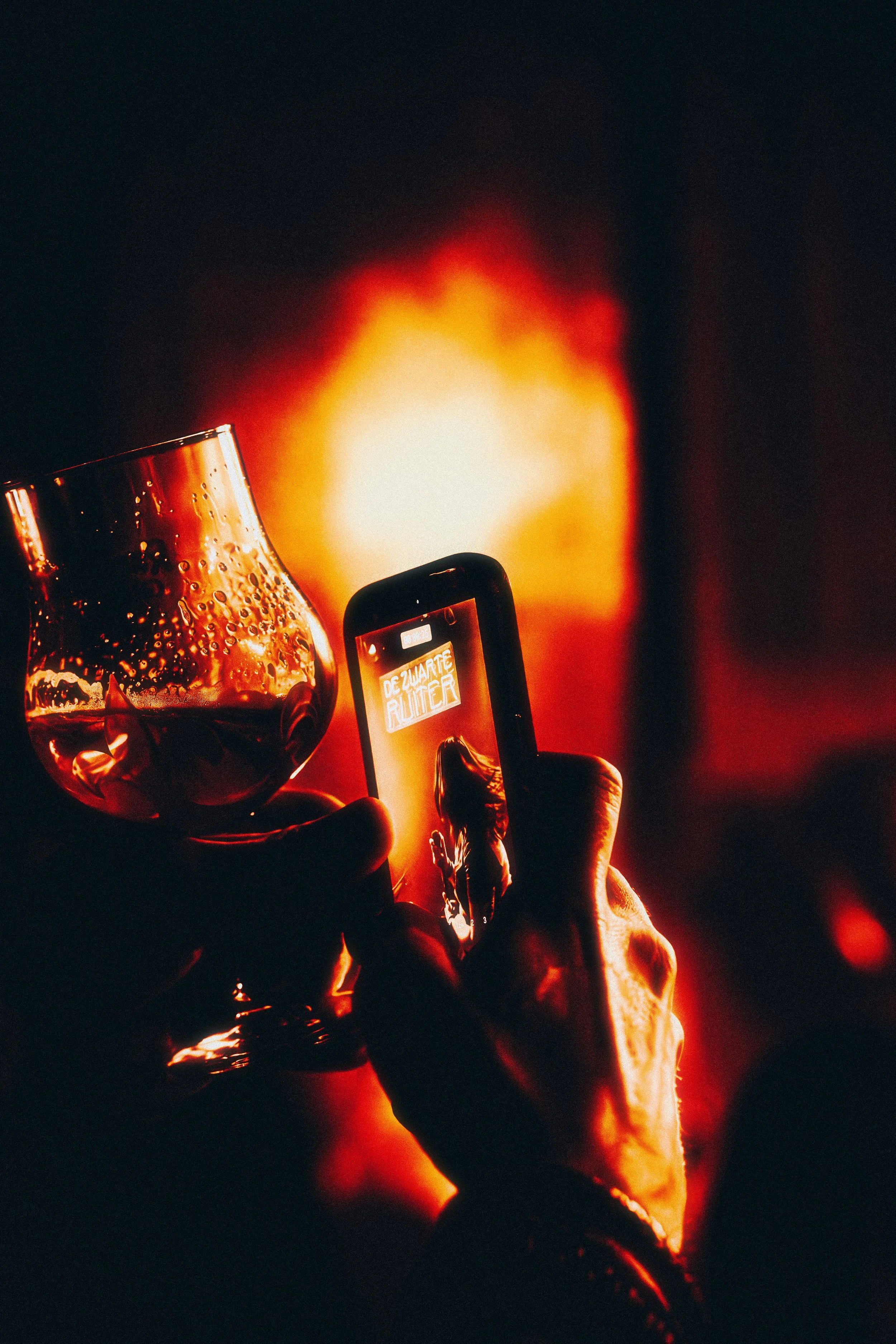 Person taking a photo with a smartphone of a wine glass filled with a drink, with a bright orange light in the background.