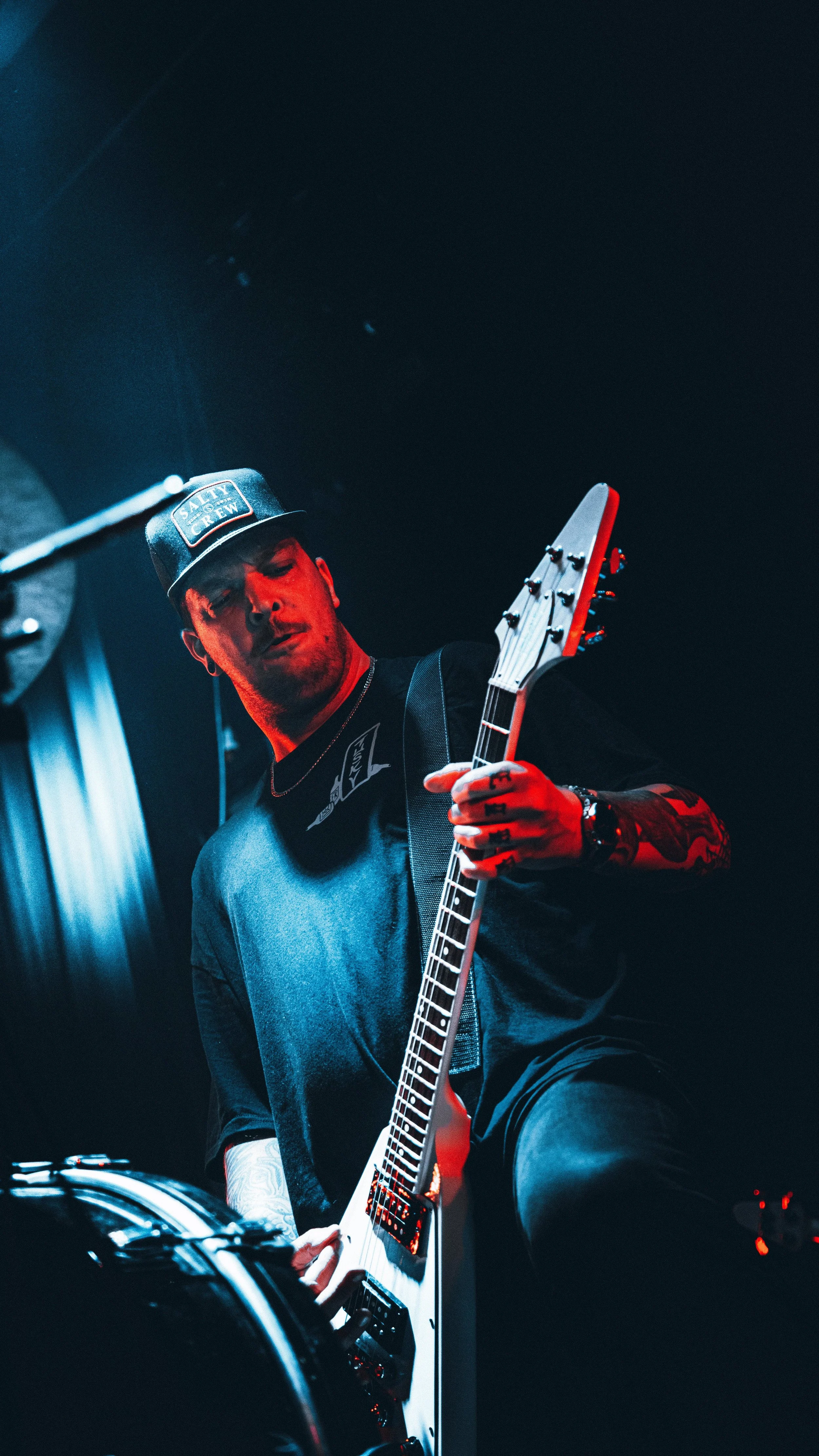 A male guitarist playing an electric guitar on stage, wearing a black T-shirt and a black cap with a logo, with red and blue stage lighting.